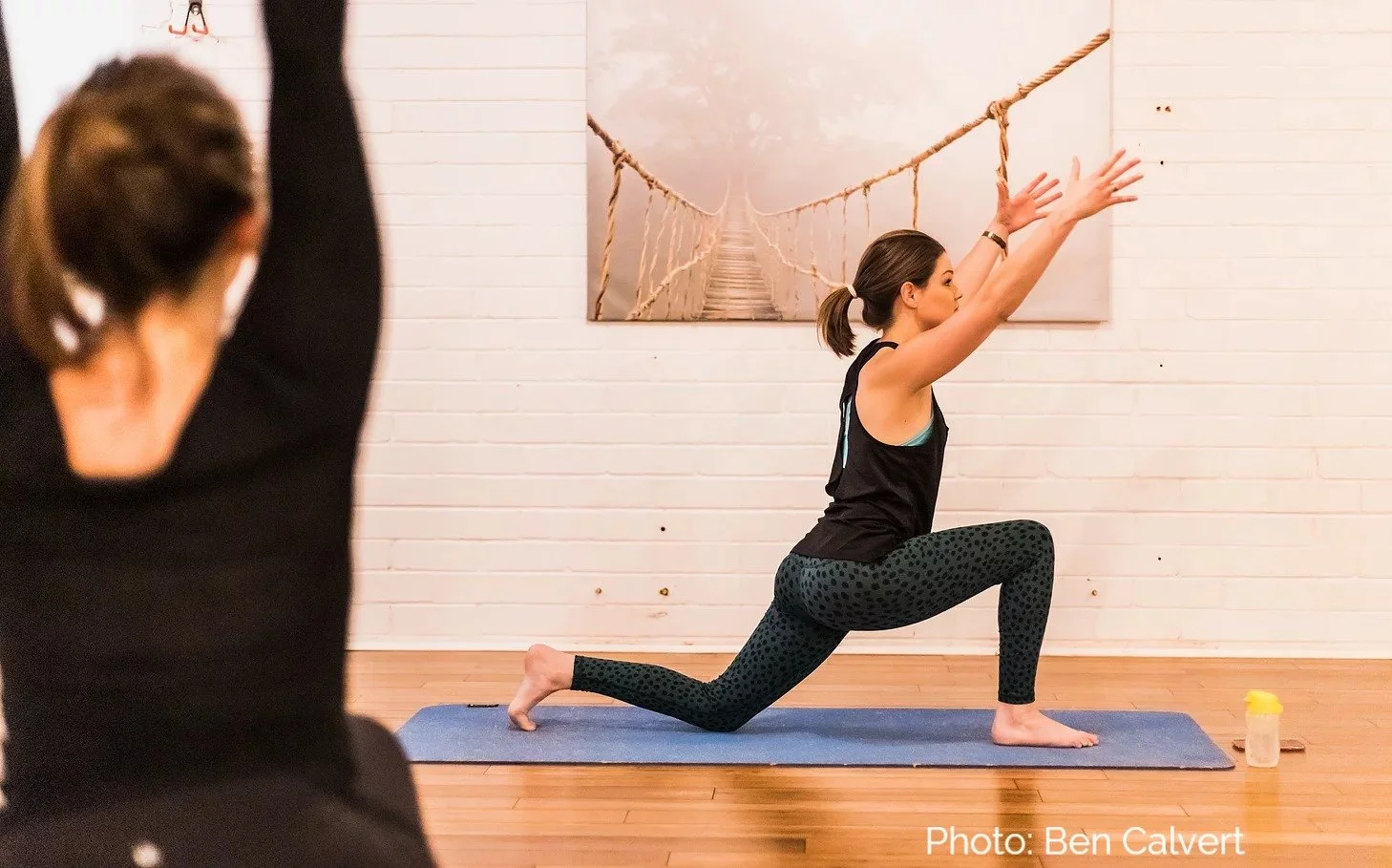 Woman practicing a yoga lunge stretch on a mat in a studio while an instructor demonstrates in the foreground