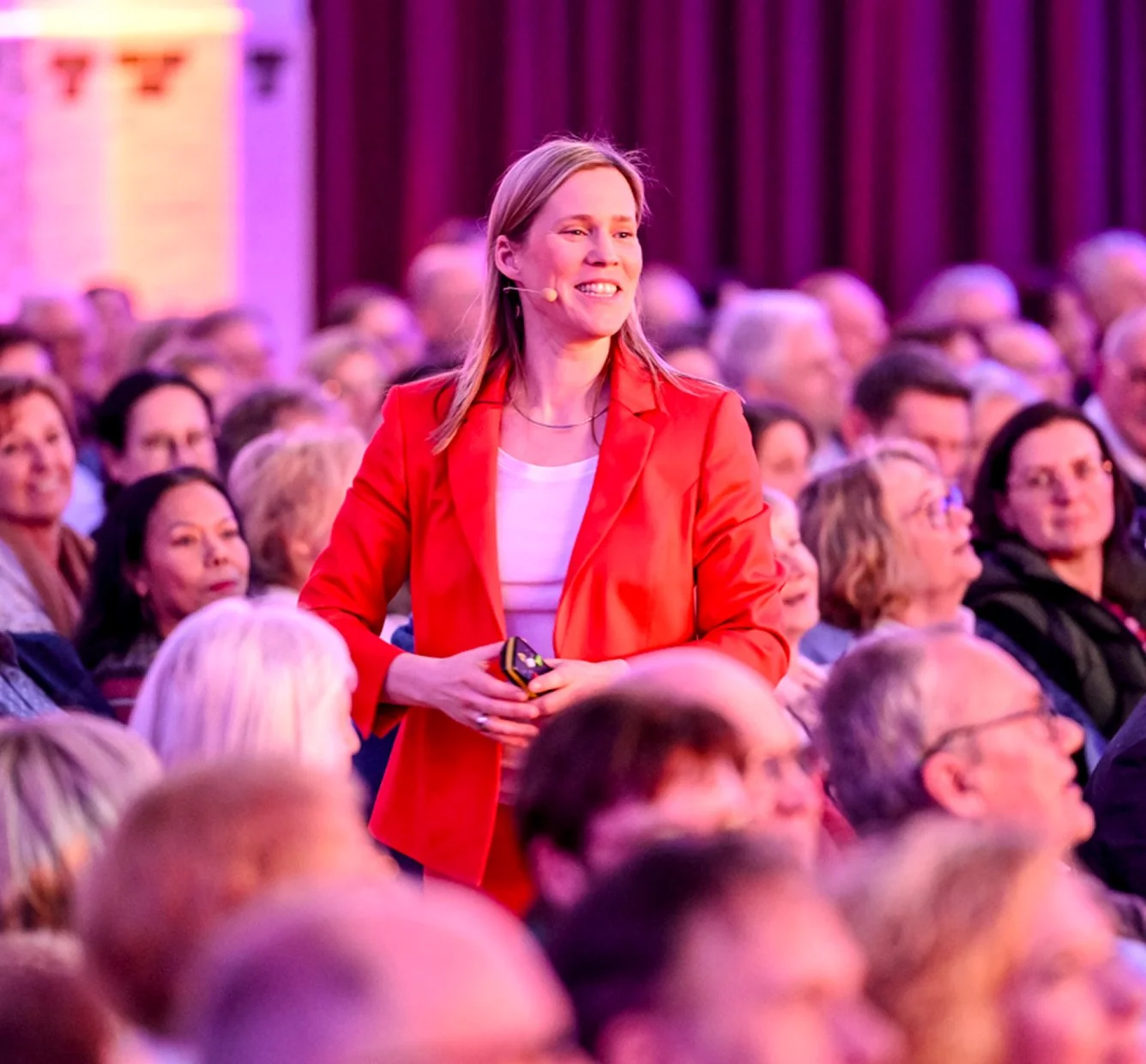 Eine lächelnde Frau in rotem Blazer bei einer Versammlung, umgeben von einem Publikum in einem großen Saal mit roten Vorhängen im Hintergrund.