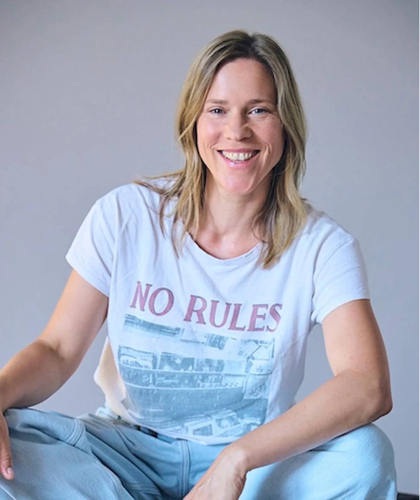 Lächelnde Frau mit Sandfarbenem, mittellangem Haar, trägt ein weißes T-Shirt mit 'NO RULES'-Aufdruck, sitzt auf dem Boden vor einem neutral grauen Hintergrund.