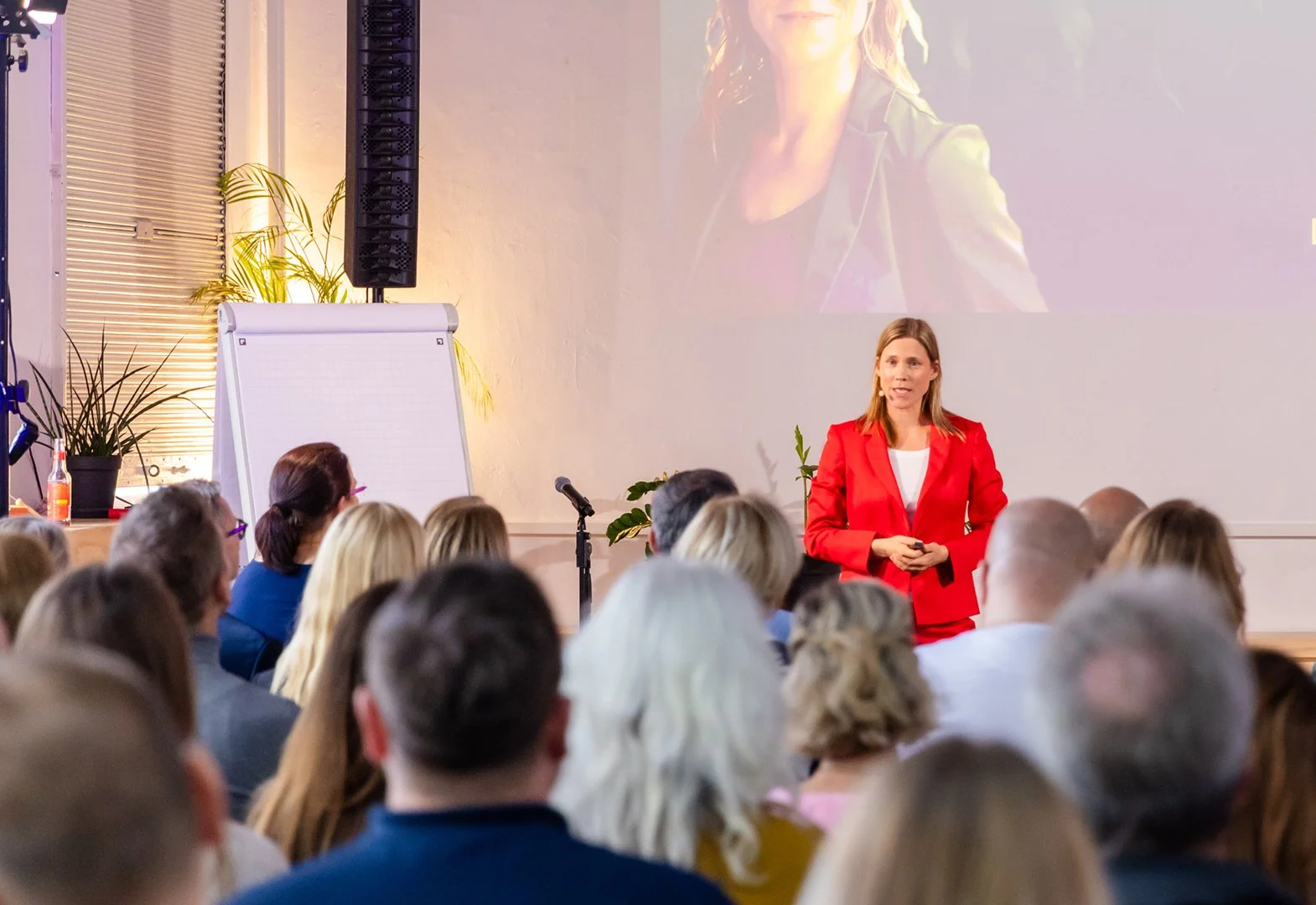 Eine Frau in rotem Blazer hält eine Präsentation vor einem Publikum in einem Seminarraum, mit Whiteboard und Pflanzen im Hintergrund.
