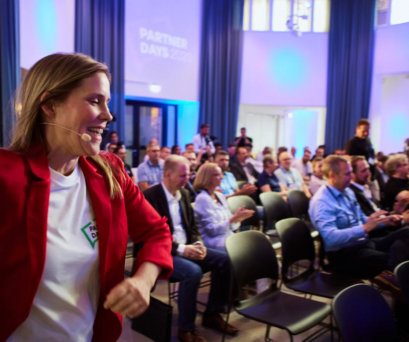 Eine lächelnde Frau mit einem Headset und rotem Mantel steht vor einer Gruppe von Menschen in einem Konferenzraum. Die Gruppe sitzt und schaut zum Vortrag. Der Raum ist gut beleuchtet mit blauen und lila Lichteffekten.