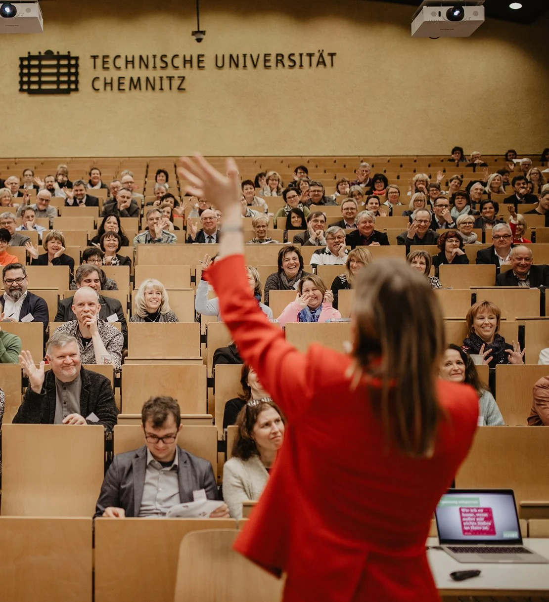Ein Redner in rotem Anzug spricht vor einer Sitzenden Menschenmenge in einem Hörsaal der Technischen Universität Chemnitz, Deutschland.