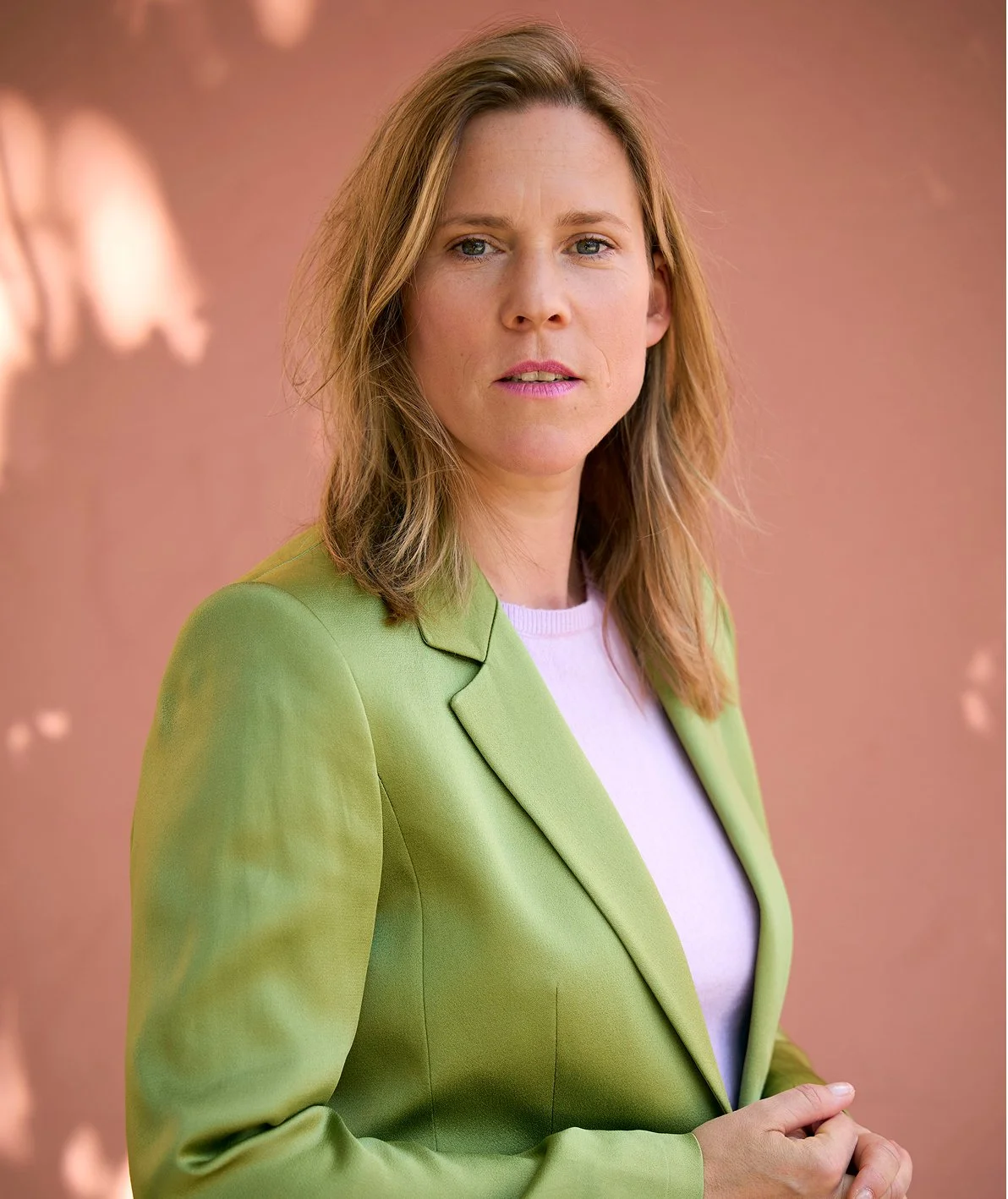 Porträt einer Frau mit blonden Haaren, blauen Augen, in einem hellgrünen Blazer und einem weißen Shirt, vor einem hellbraunen Hintergrund.