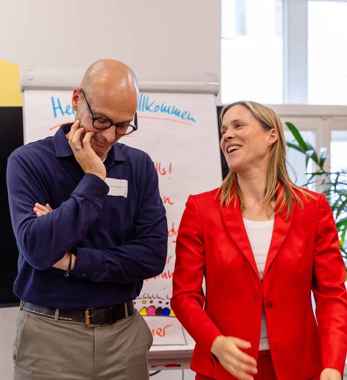 Zwei lächelnde Menschen, ein Mann und eine Frau, stehen vor einem Flipchart mit deutschem Text, scheinbar bei einem Meeting oder Workshop.