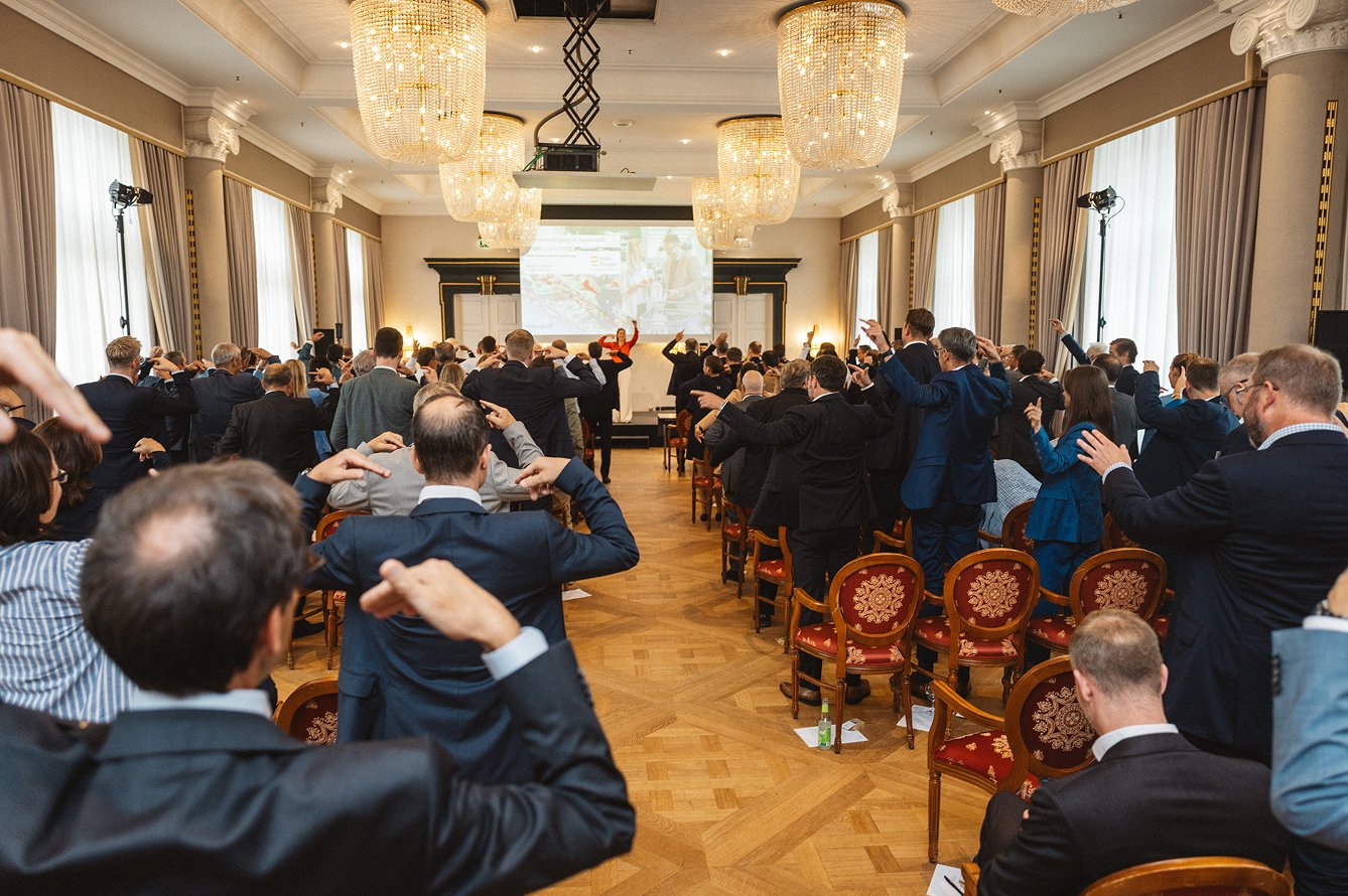 Eine große Gruppe von Menschen in einem eleganten Veranstaltungsraum nehmen an einer Zeremonie oder einem Treffen teil, bei dem alle nach vorne ihre rechte Hand auf die Stirn legen.