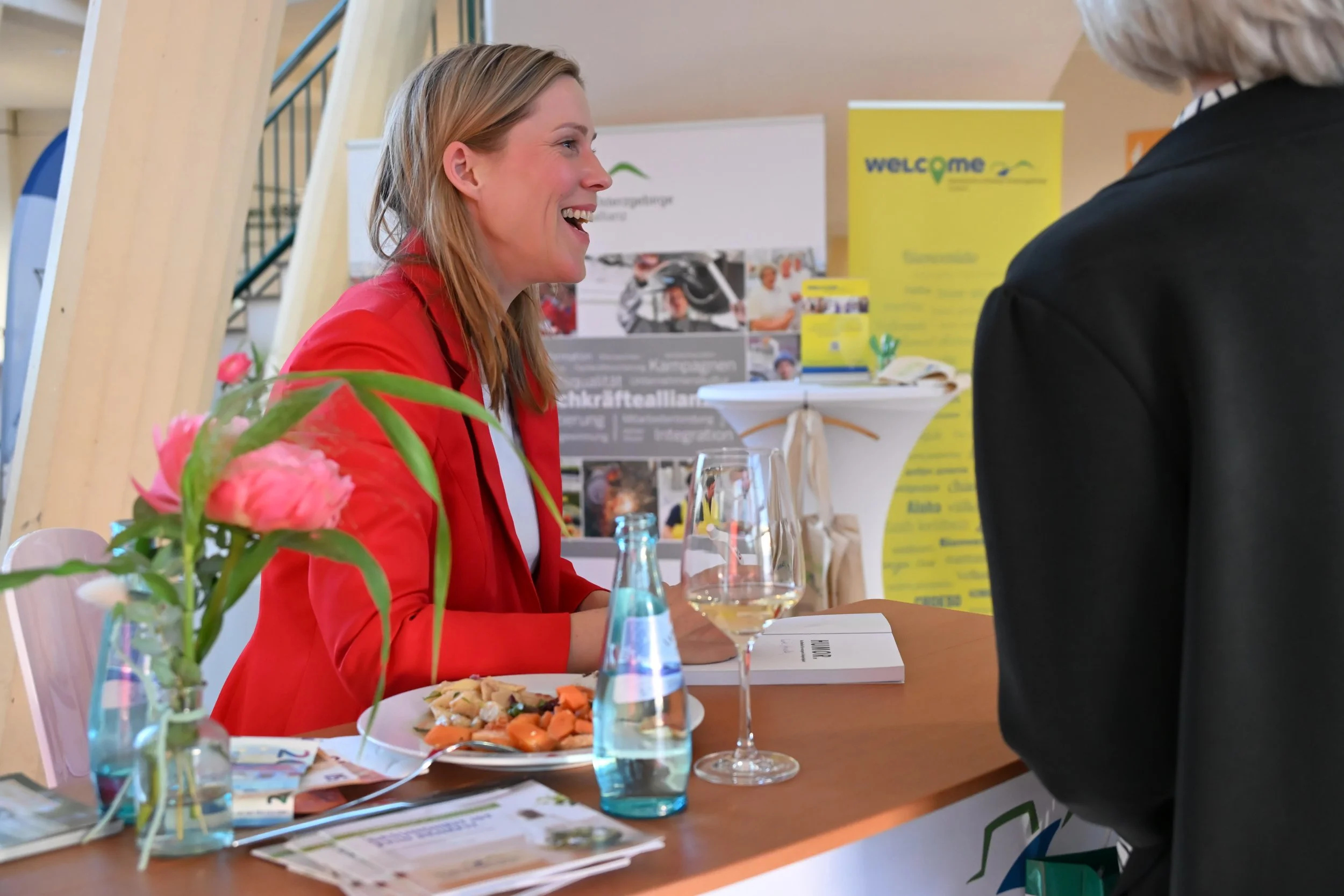 Eine Frau in roten Jacke spricht mit einer anderen Person bei einem Treffen. Auf dem Tisch stehen Getränke und eine Platte mit Essen, im Hintergrund Banner und Infomaterialien.