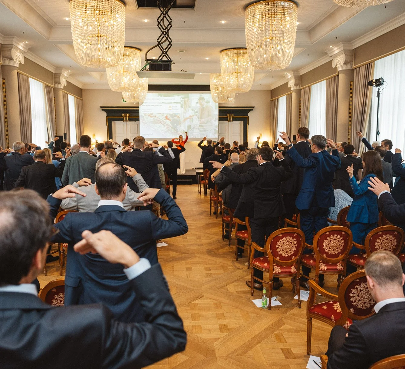 Ein Konferenzraum mit vielen Geschäftsleuten, die an einem Seminar teilnehmen und einer Sprecherin auf der Bühne. Die Teilnehmer sitzen in Stühlen mit rotem Sitzpolster, die an den Wänden hängen, und machen Handzeichen.