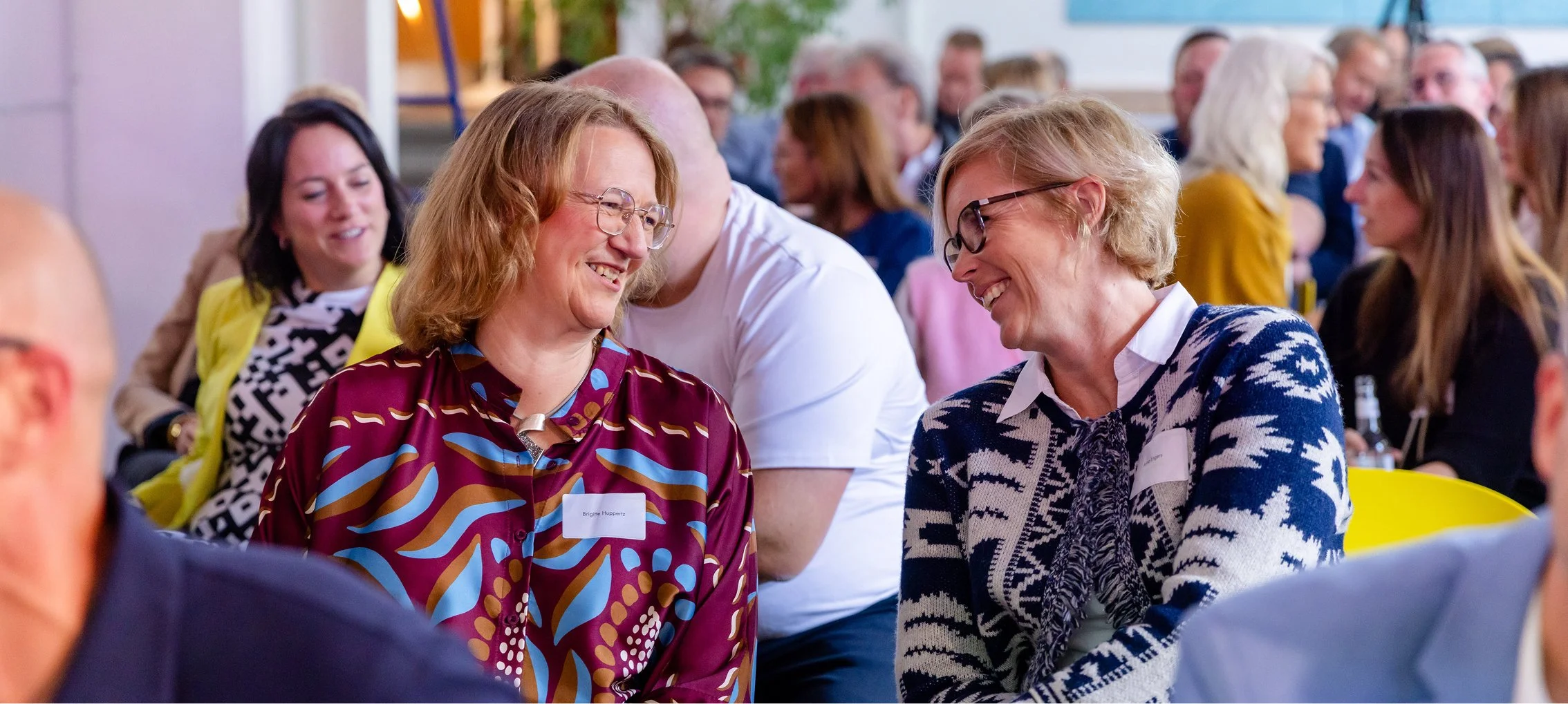 Zwei Frauen im Gespräch, lachend und freundlich, bei einer Veranstaltung mit vielen Menschen im Hintergrund.