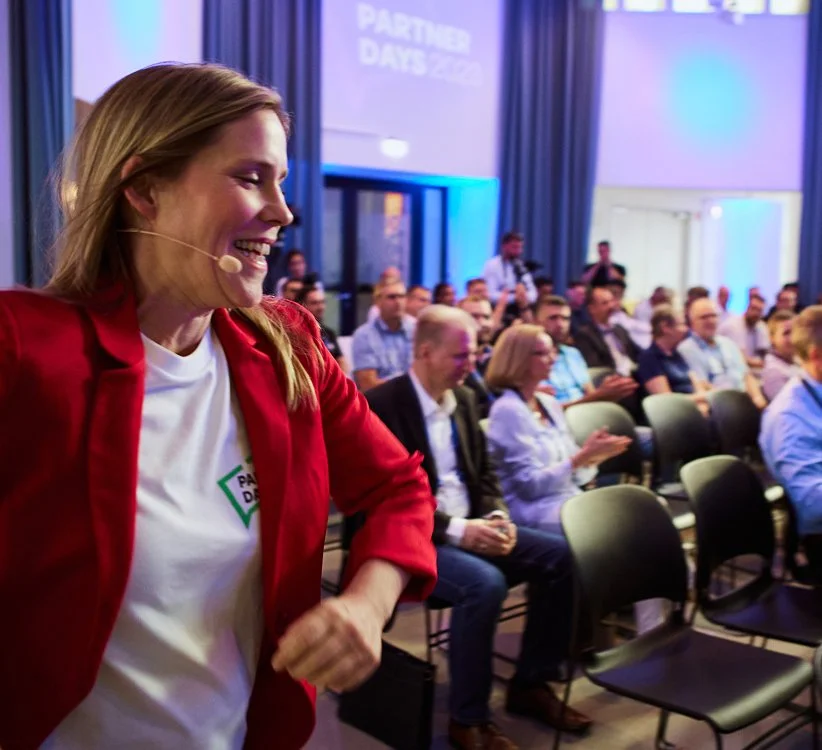 Eine lachende Frau mit einem Headset auf einem roten Blazer bei einer Konferenz, im Hintergrund sitzen ein Publikum und eine große Leinwand.