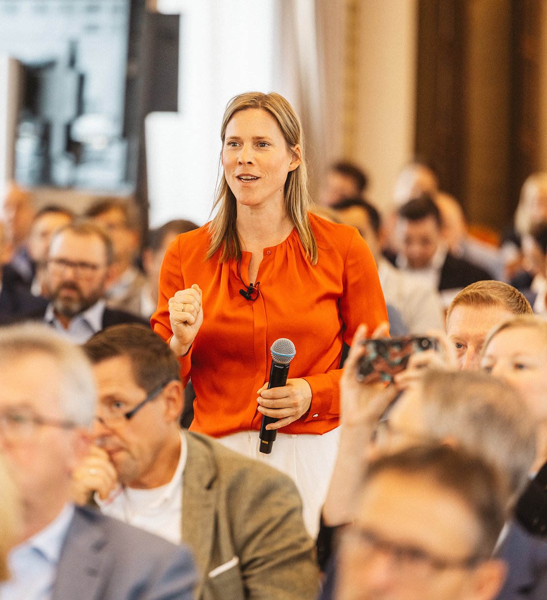 Eine Frau in rotem Hemd hält ein Mikrofon während einer Podiumsdiskussion vor einem Publikum.