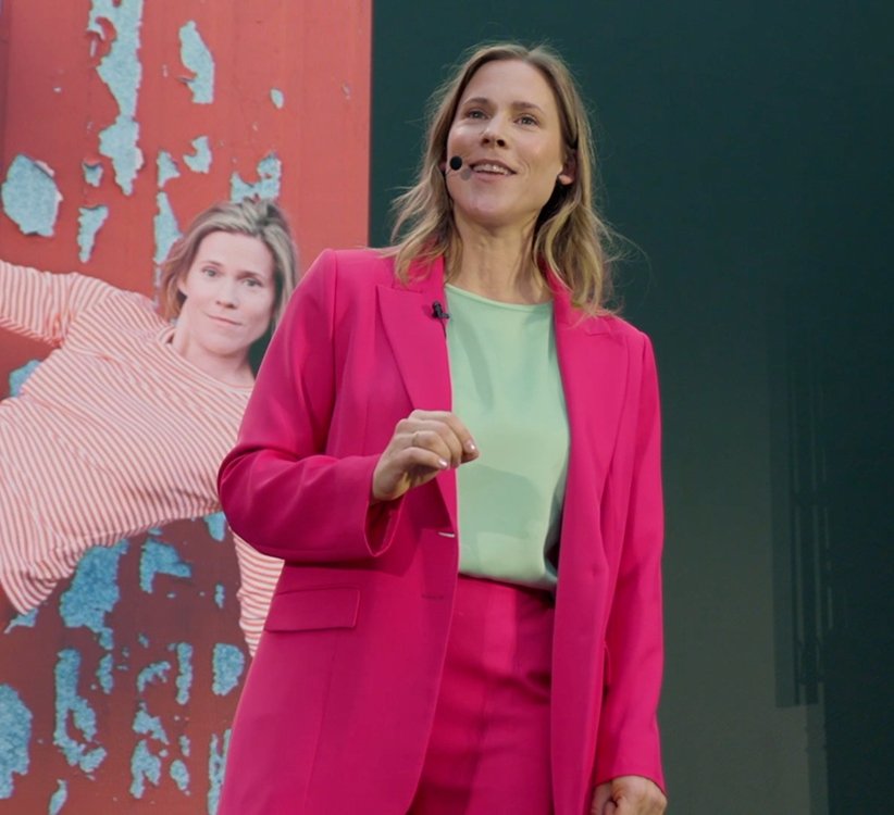 Frau in pinkem Anzug spricht auf einer Bühne, im Hintergrund ein Poster mit einem lächelnden Gesicht