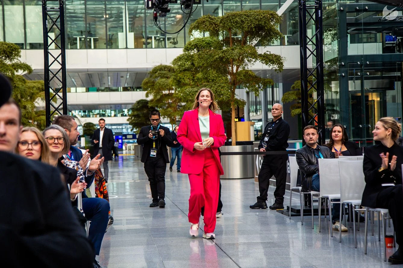 Eine Frau in einem pinken Anzug läuft durch eine moderne Kongresshalle, umgeben von Menschen, die im Publikum sitzen und applaudieren.