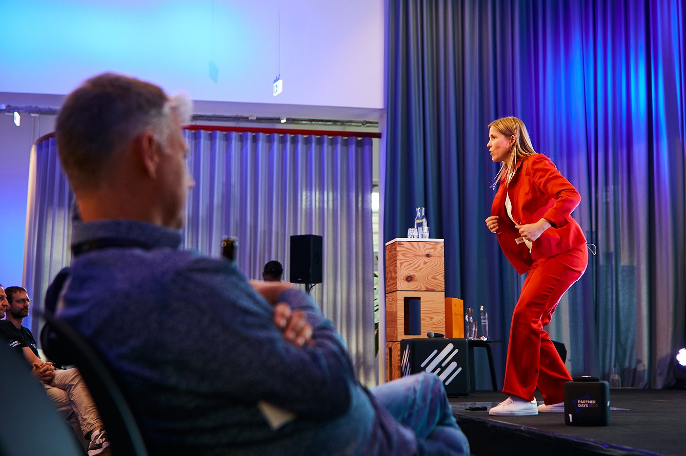 Eine Frau in rotem Anzug auf einer Bühne bei einer Präsentation, im Hintergrund lila und blaue Beleuchtung, Publikum im Saal.