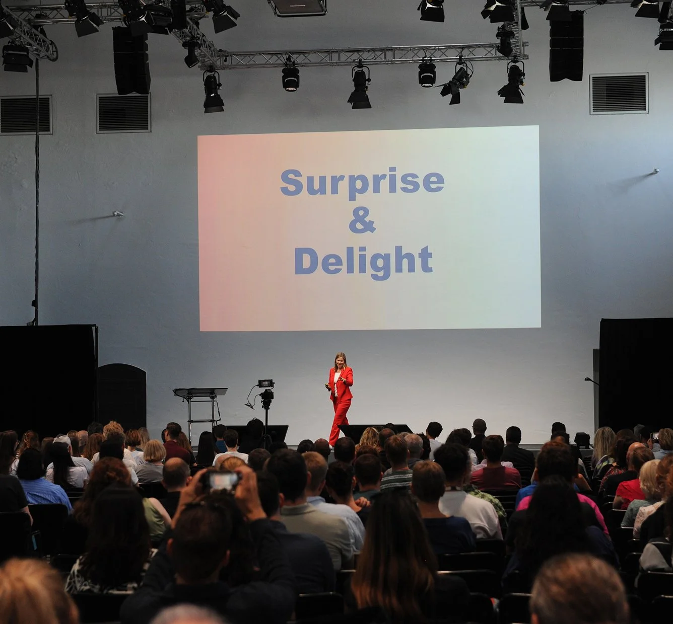 Eine Präsentation in einem großen Saal mit einer Frau in rotem Anzug auf der Bühne vor einem Publikum. Auf der Leinwand steht "Surprise & Delight".