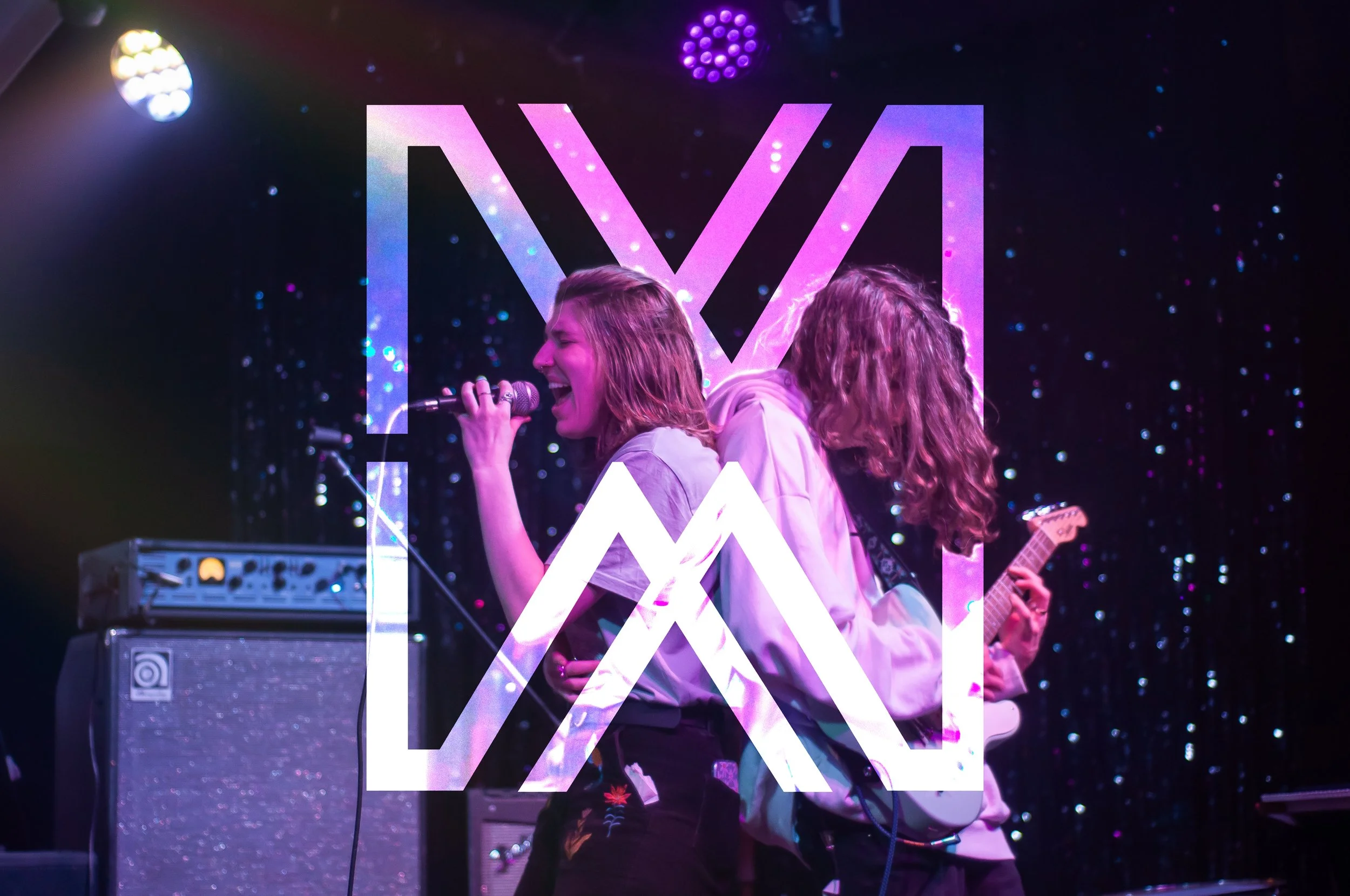 A woman singing into a microphone back to back with a long-haired man playing guitar, semi-transparent overlay of the logo 'MW'.