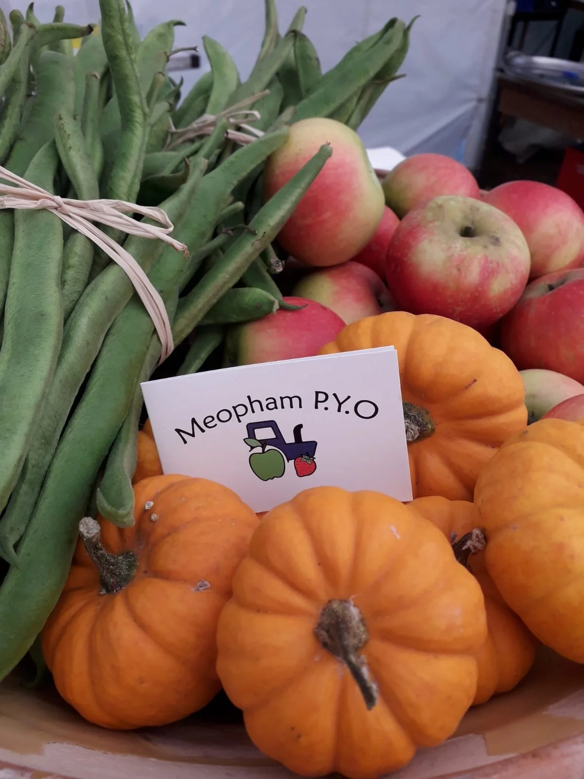 A basket of small orange pumpkins, green beans, and red apples with a sign that reads 'Meopham P.Y.O', indicating pick-your-own farm produce.