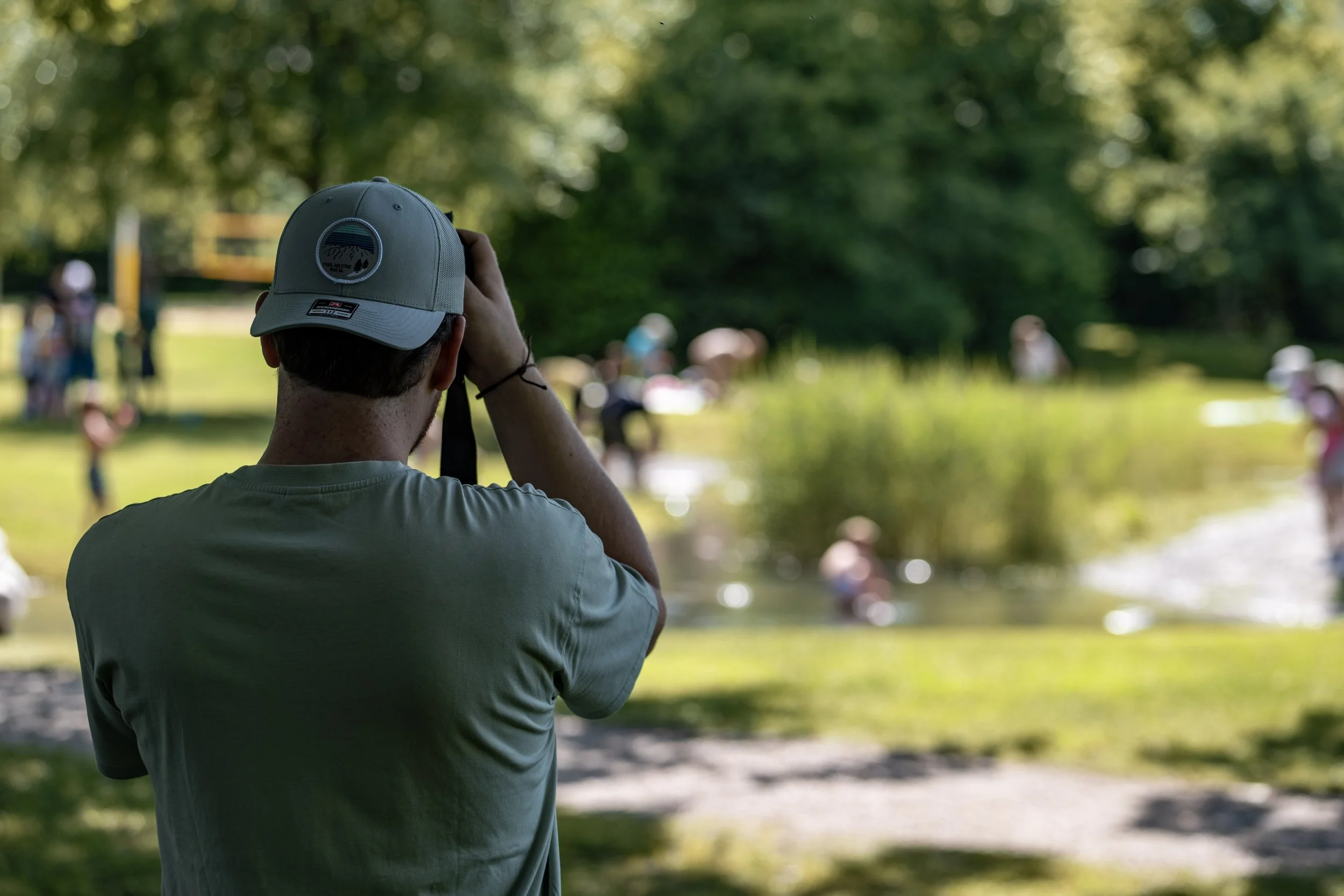 Mensch mit grauem Baseball-Cap fotografiert eine Szene im Park, im Hintergrund spielen Kinder im Wasser.