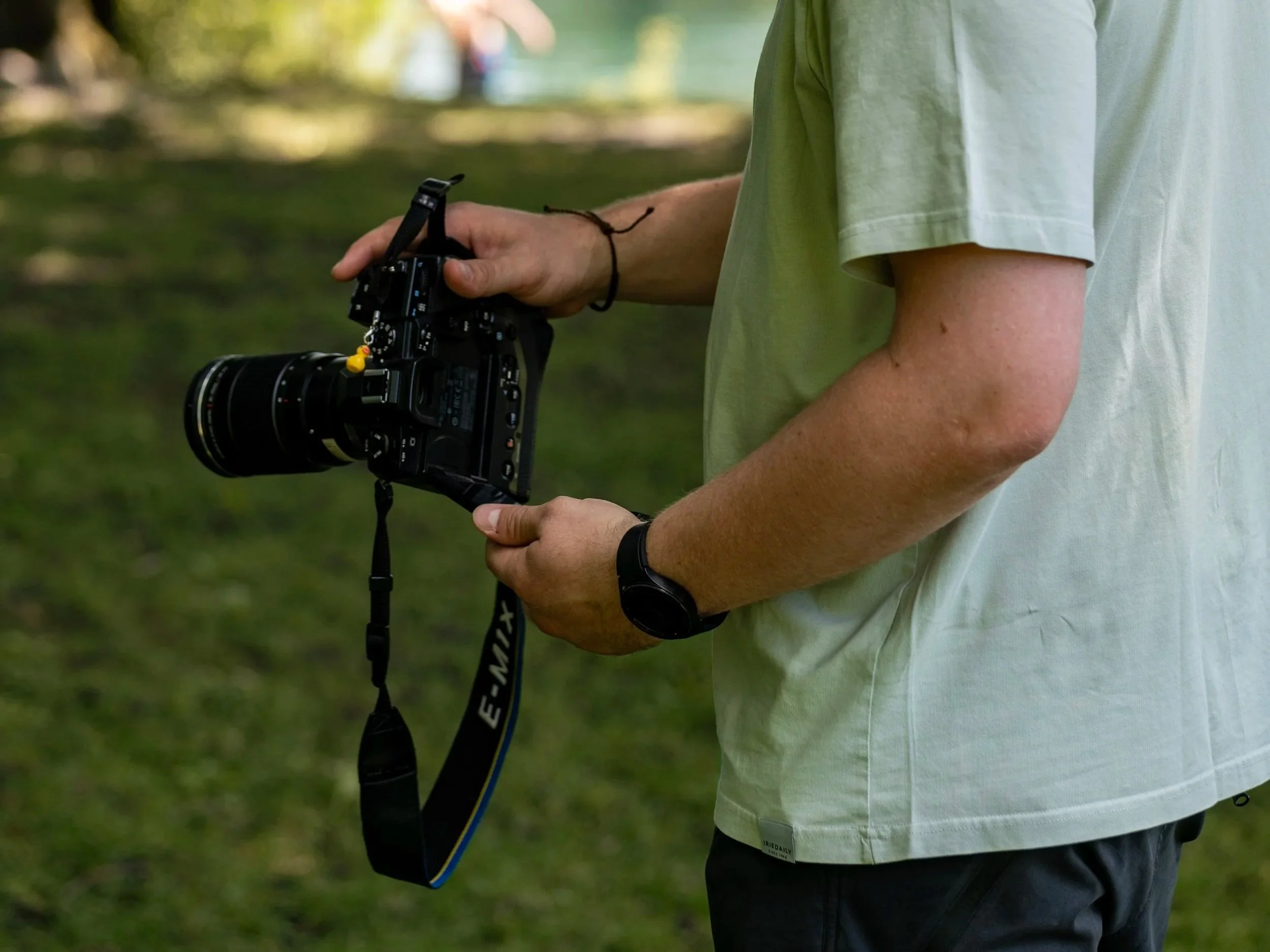 Person in hellem T-Shirt hält professionelle Digitalkamera mit großem Objektiv im Freien, vermutlich bei einer Fotosession oder im Park.