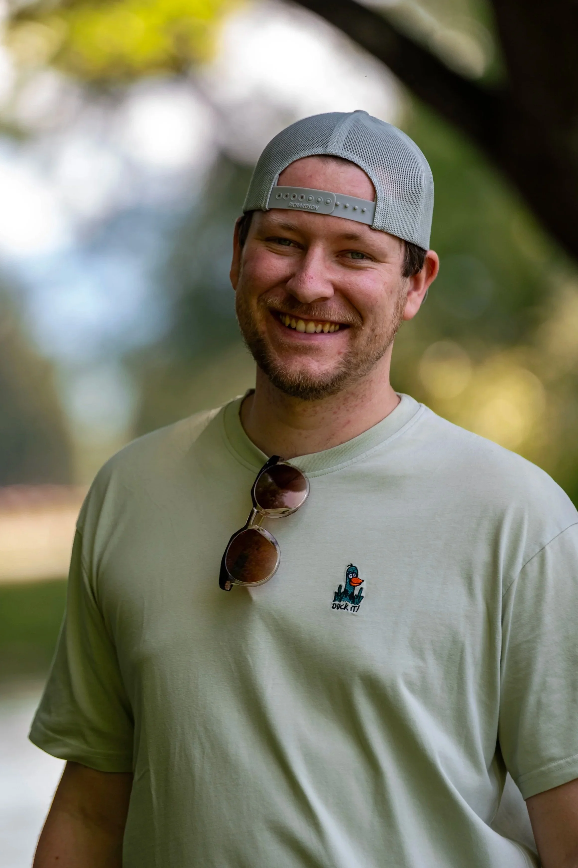 Nahaufnahme eines lächelnden Mannes im Freien, trägt ein graues Baseballcap, eine hellgrüne T-Shirt mit einem Duck-Logo und Sonnenbrille. Hintergrund unscharf mit Bäumen und Himmel.
