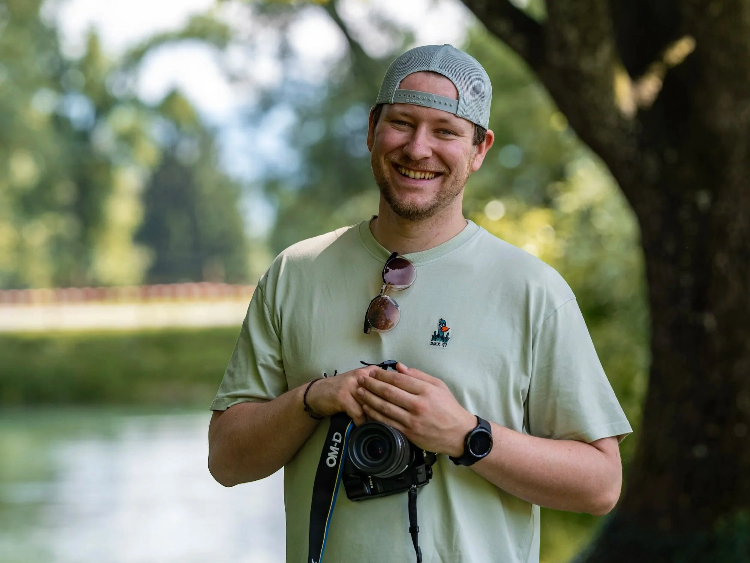 Ein lachender Mann trägt ein graues Baseball-Cap, eine Sonnenbrille, ein weißes T-Shirt mit einer Ente und hält eine Kamera in den Händen in einem Park mit Bäumen im Hintergrund.