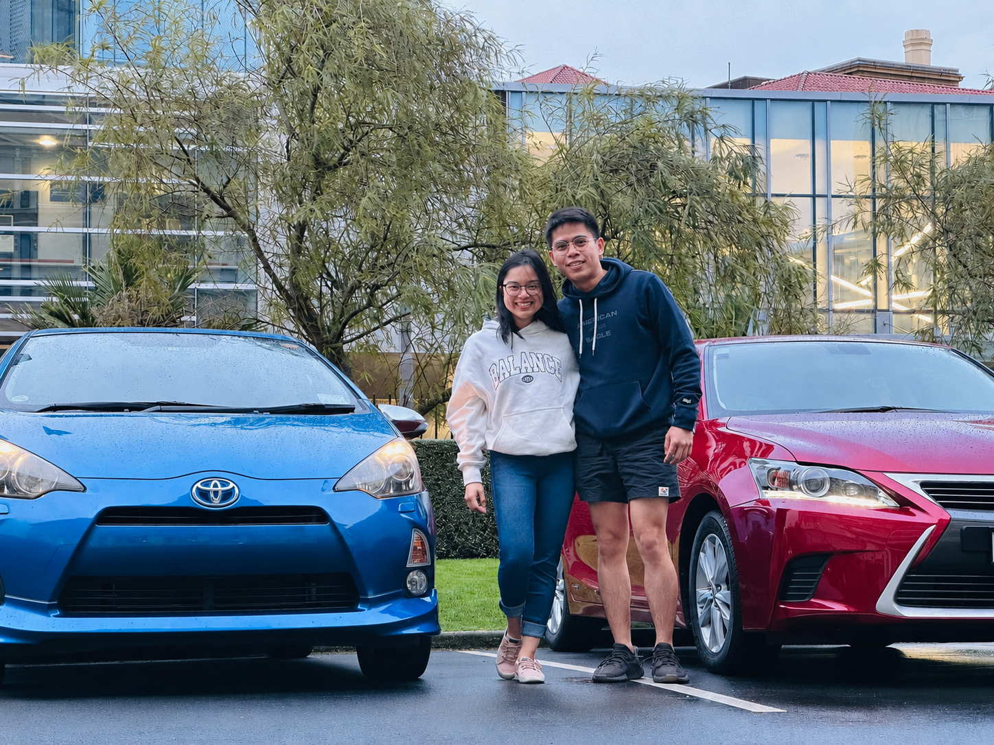 A young woman and a young man are standing arm in arm between a blue Toyota and a red Lexus in a parking lot with wet pavement and rainy weather. They are smiling and looking at the camera, with a background of trees and modern office buildings.