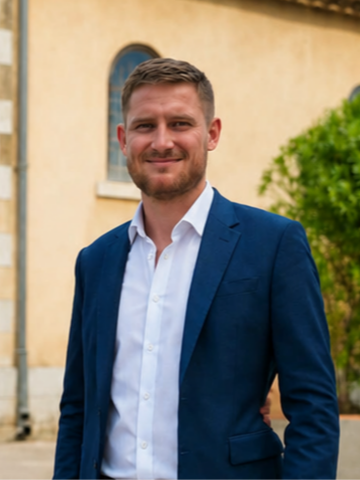 Un homme souriant portant un costume bleu clair et une chemise blanche, debout devant un bâtiment avec des fenêtres et un arbre vert en arrière-plan.