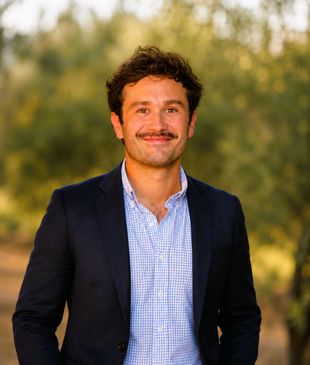 Un homme souriant en costume dans un paysage naturel avec des arbres en arrière-plan