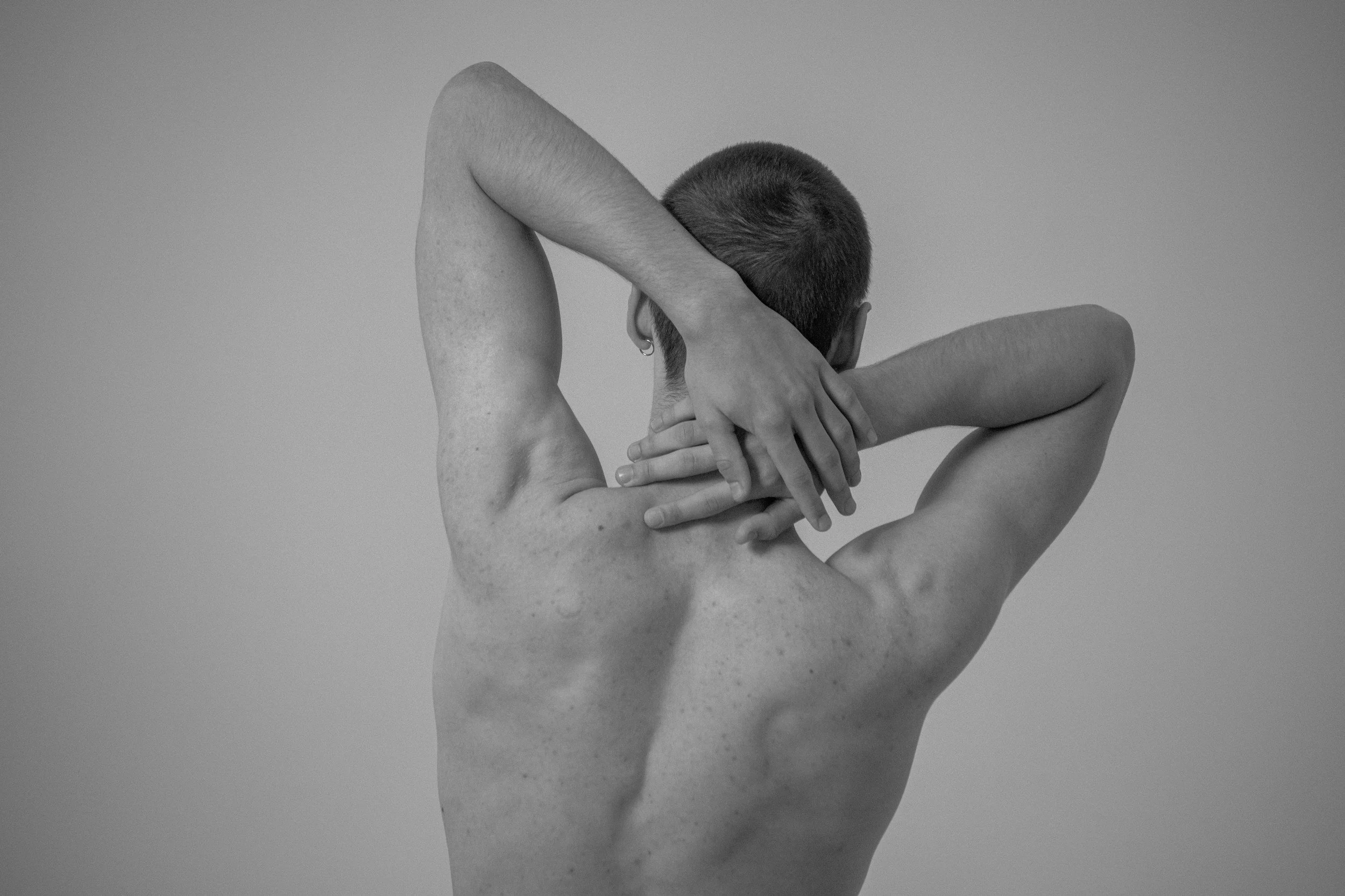 Persona con cabello corto, sin camisa, en una pose artística, con brazos cruzados detrás del cuello y las manos en la parte superior de la cabeza, en un fondo gris