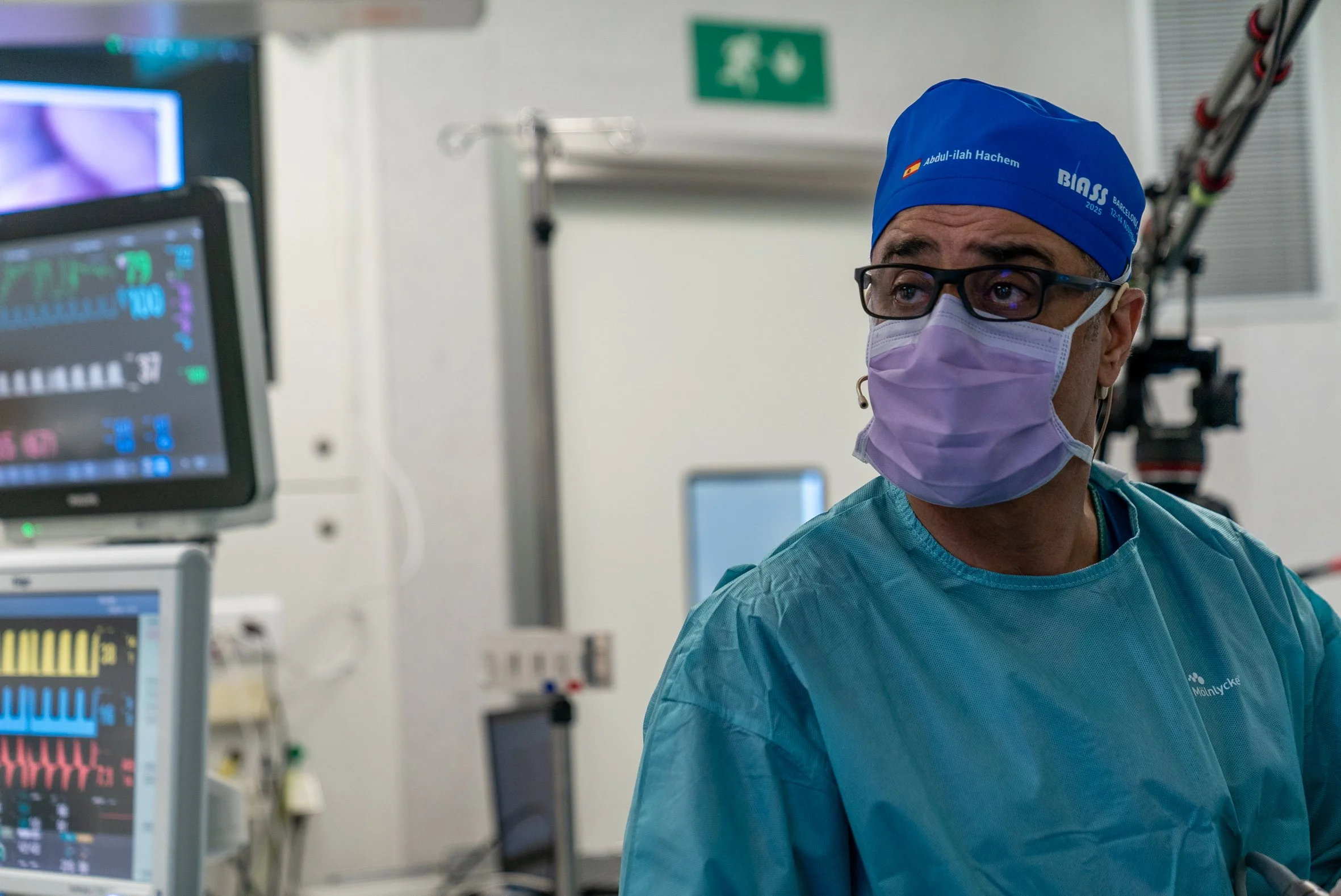 A healthcare professional wearing glasses, a purple face mask, and a blue cap with text, working in a medical setting with monitoring equipment in the background. Dr. Hachem, shoulder surgeon, Barcelona,  BIASS, Spain, cirugia de hombro, teknon