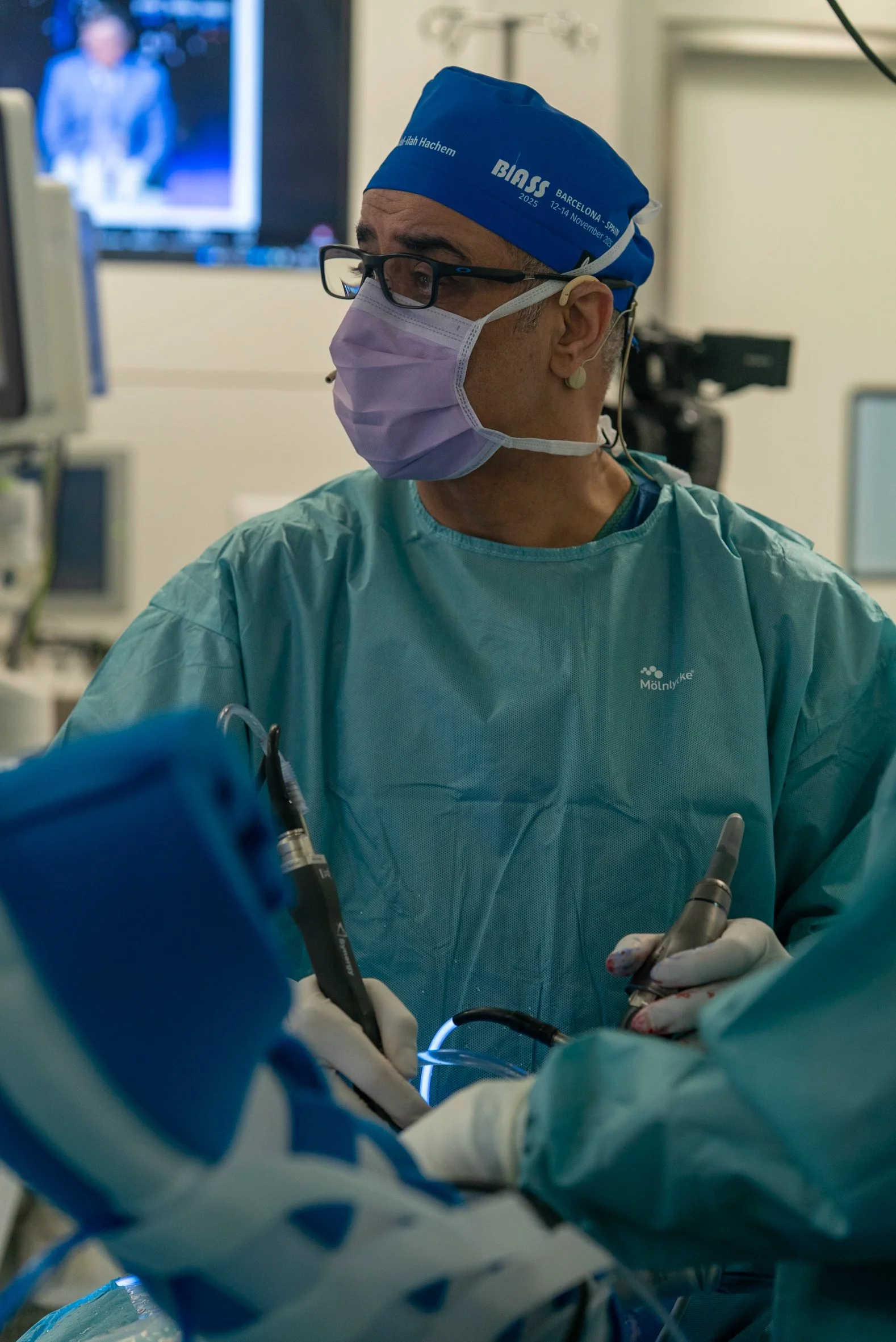 A surgeon wearing glasses, a face mask, and a blue surgical cap, operating in an operating room. In the background, there is a monitor with a blurred image. Dr. Hachem, shoulder surgeon, Barcelona,  BIASS, Spain, cirugia de hombro, teknon