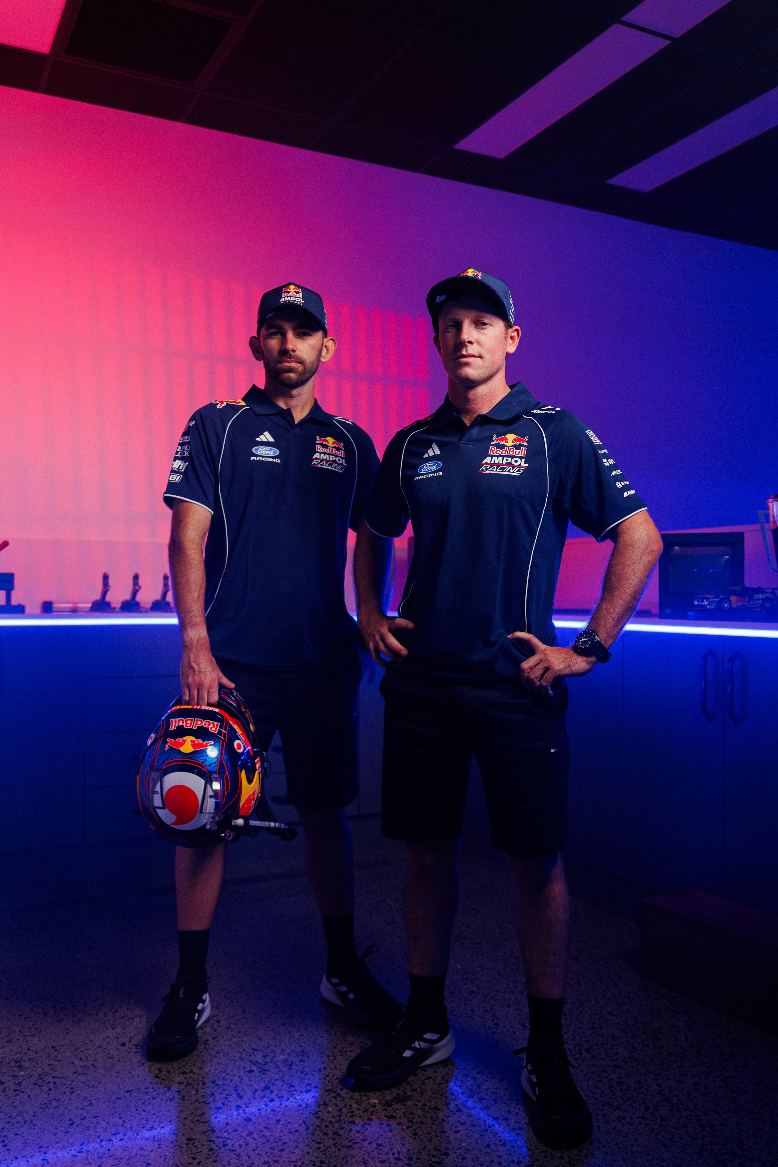 Two men in dark racing team uniforms standing in a neon-lit room, one holding a colorful racing helmet, with a purple and pink gradient wall in the background.