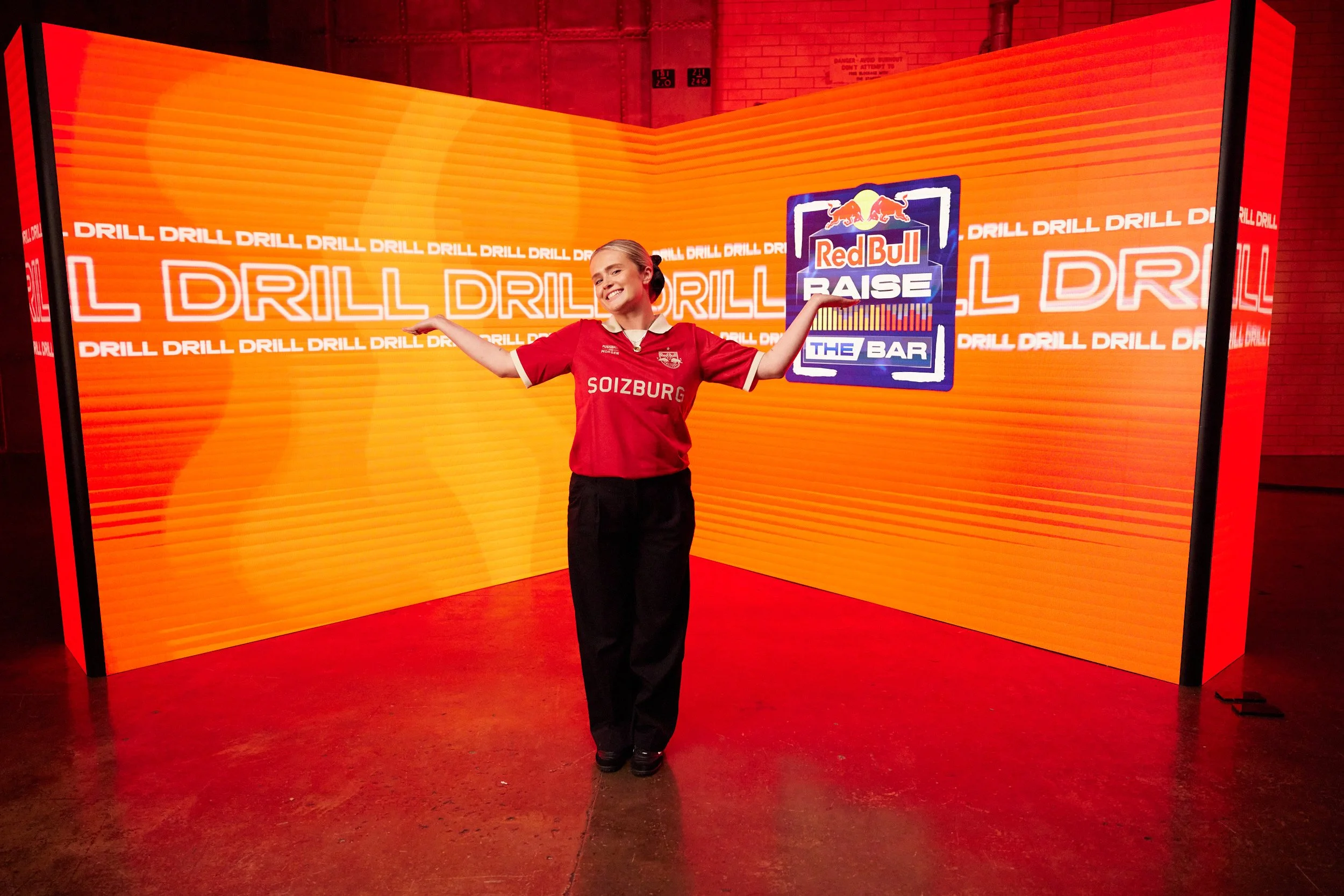 A woman in a red Red Bull racing jersey standing in front of a bright orange backdrop with the words 'DRILL' repeated and a logo for Red Bull Raise The Bar, smiling with her arms outstretched.