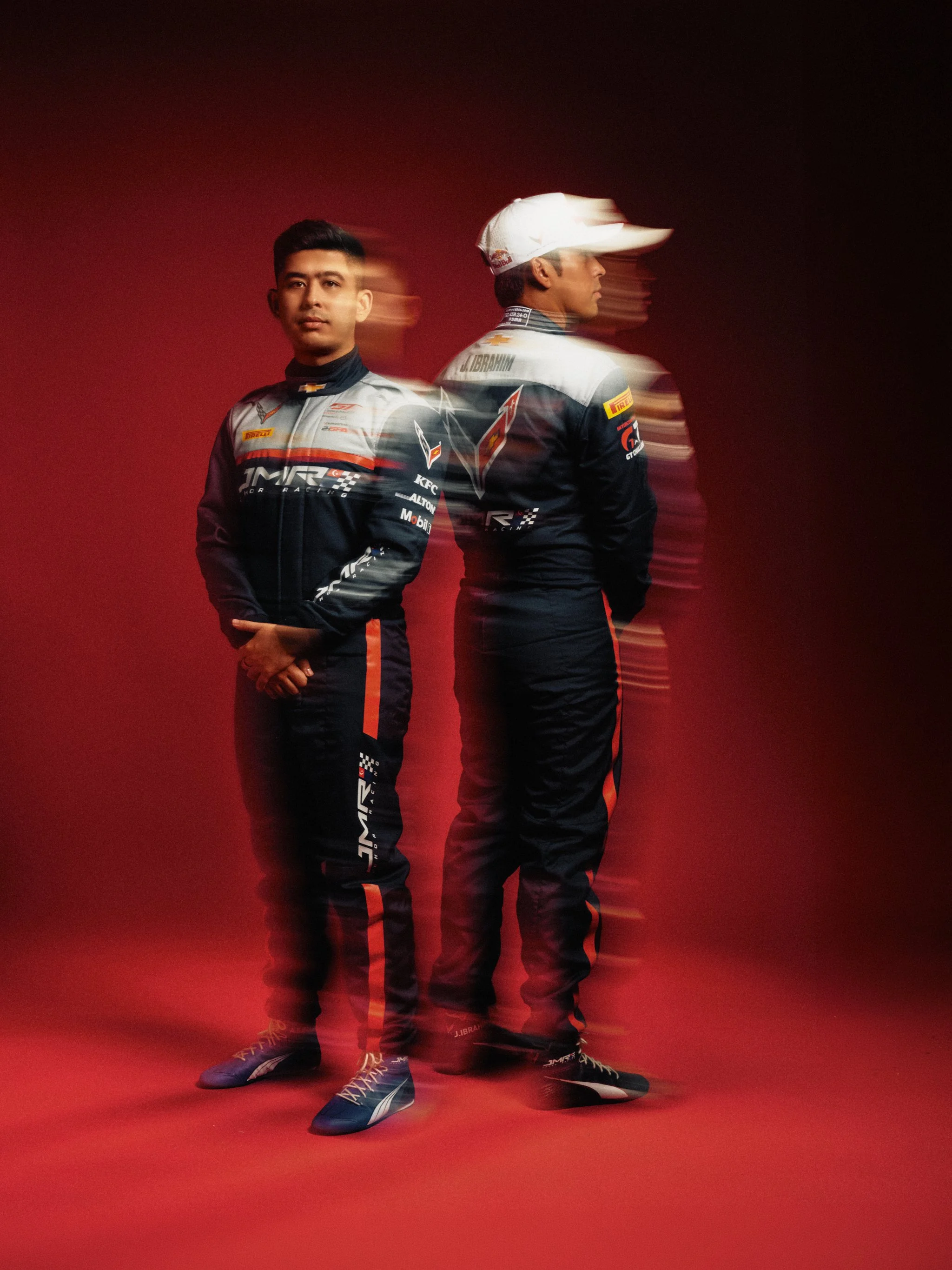 Two race car drivers wearing racing suits and shoes, standing on a red surface with a gradient red and dark background. One driver is facing forward, and the other is turned to the side, with motion blur around them.