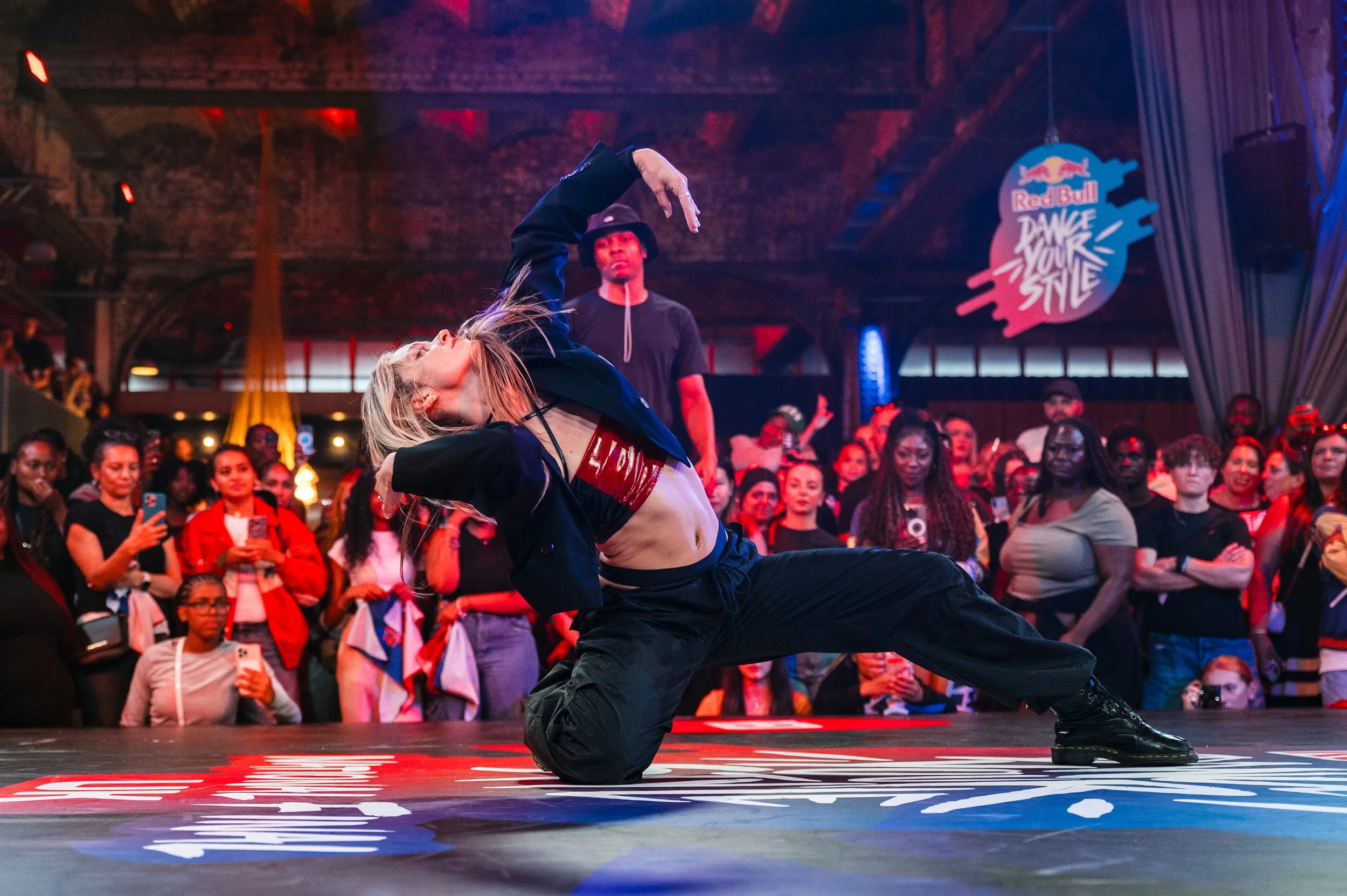 A dancer performing a dance move on stage at a Red Bull event with a crowd watching in the background. The stage has a Red Bull logo and sign that says 'Dance Your Style'.
