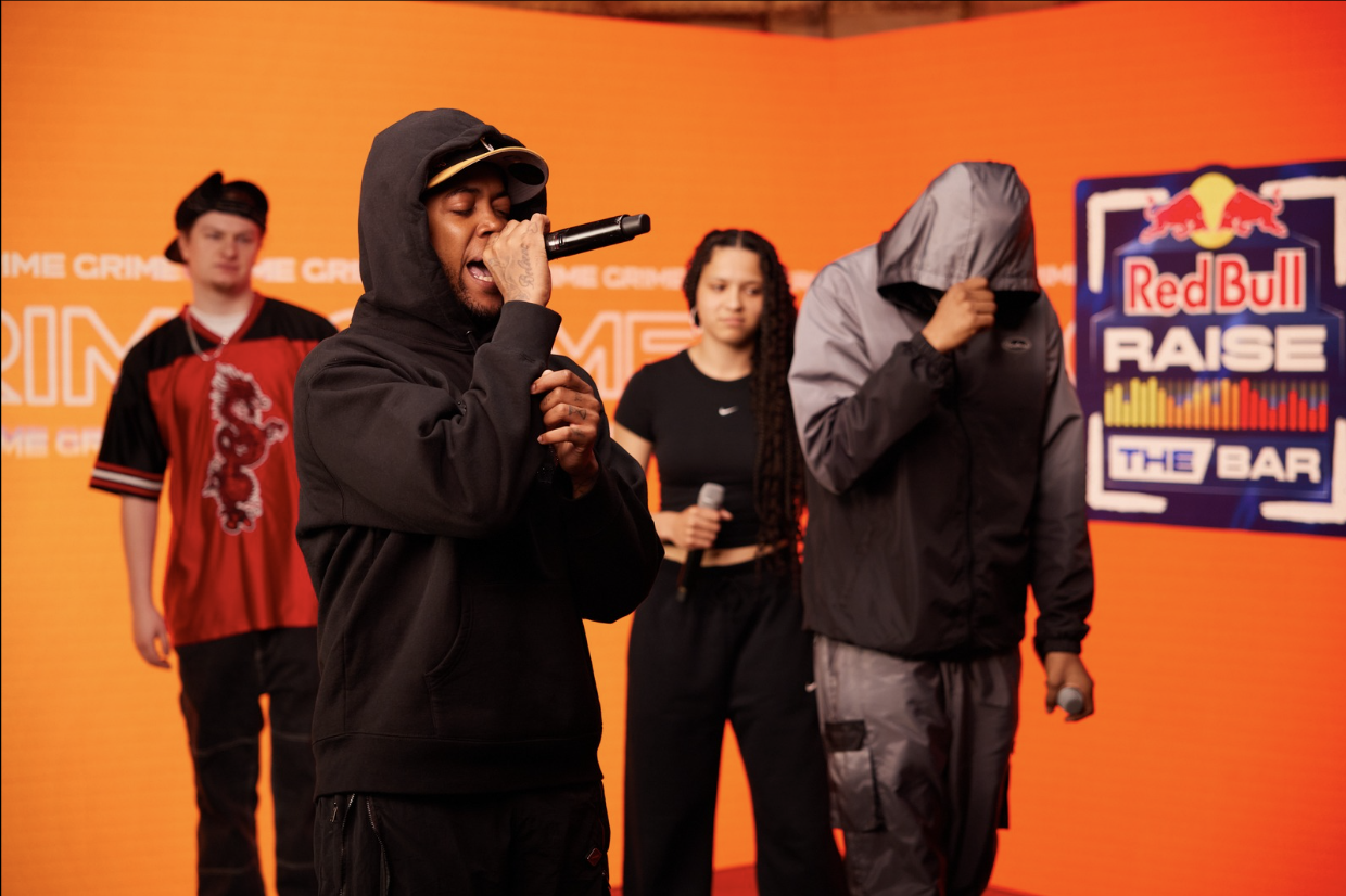Four people performing on stage with a Red Bull Raise the Bar logo in the background. The person in the foreground is singing with a microphone, wearing a black hoodie and cap. Another person beside them has their face covered by a gray hood, and the other two are in the background, one holding a microphone and the other standing with their arms crossed.