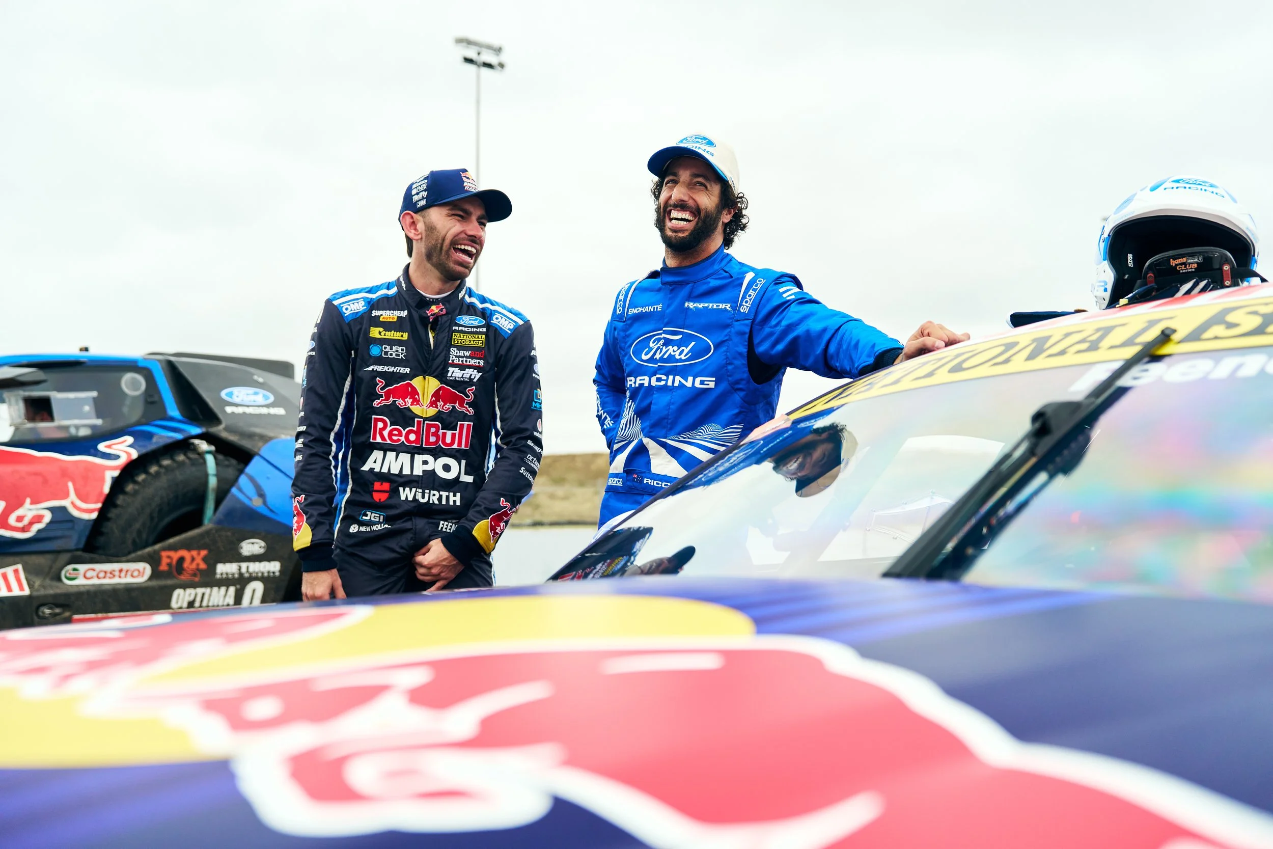 Two race car drivers with dark hair and beards standing near race cars, smiling and talking during a race event. One is wearing a black and yellow racing suit with Red Bull Racing logos, and the other is in a blue racing suit with Ford Racing branding. A race helmet is placed on one of the cars.