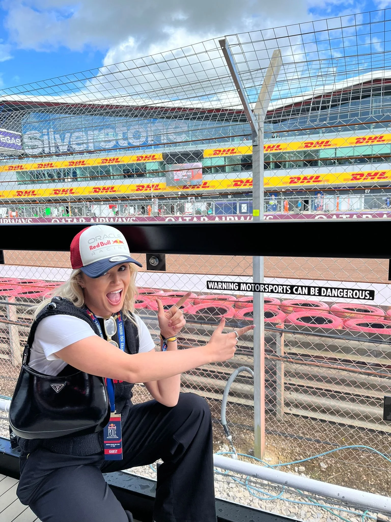 A woman in Red Bull Racing team attire smiling and pointing towards the racetrack barrier, with a racing circuit and grandstand in the background.