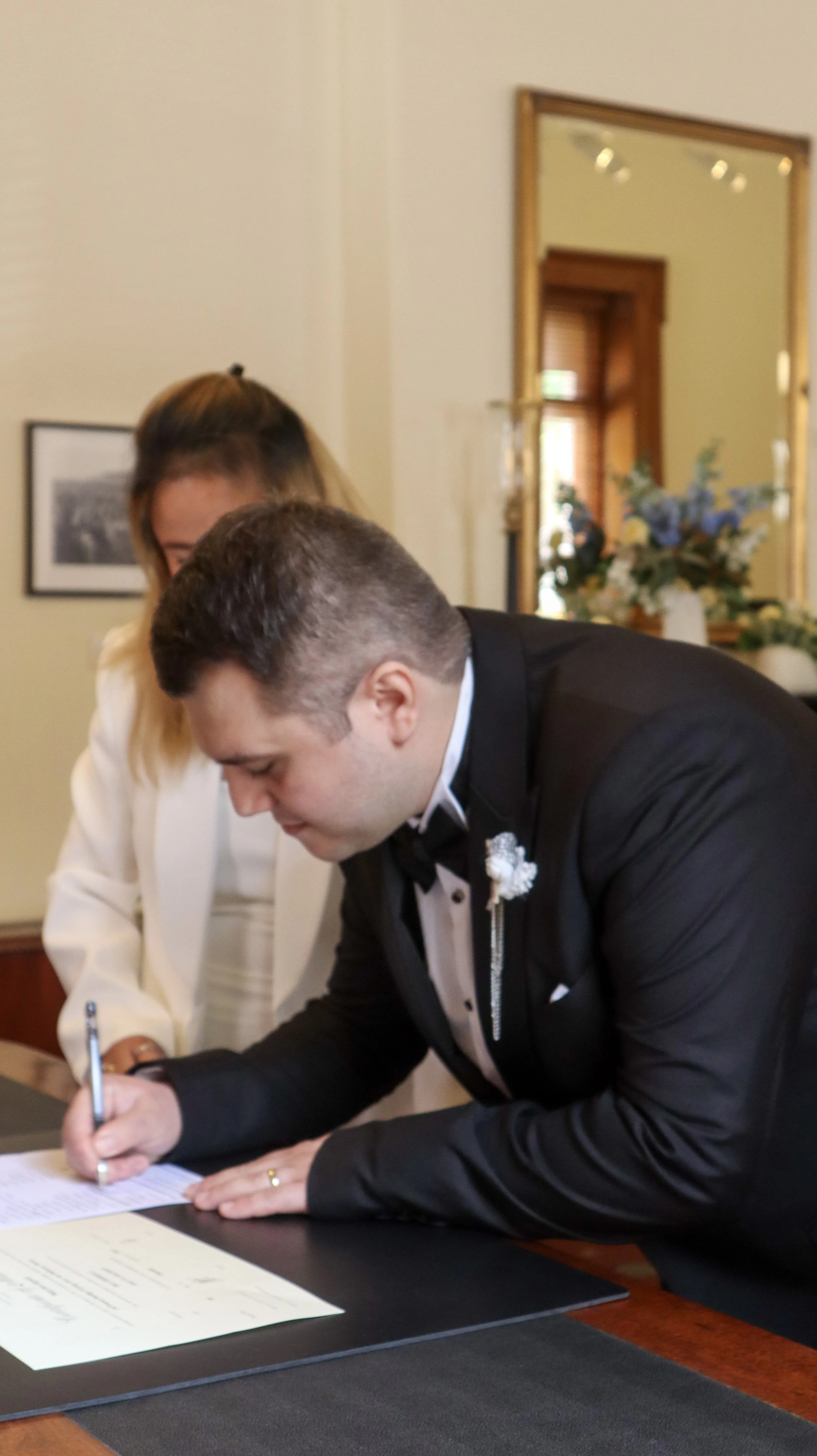Man and woman signing documents at a formal event, with a mirror and flower arrangement in the background.