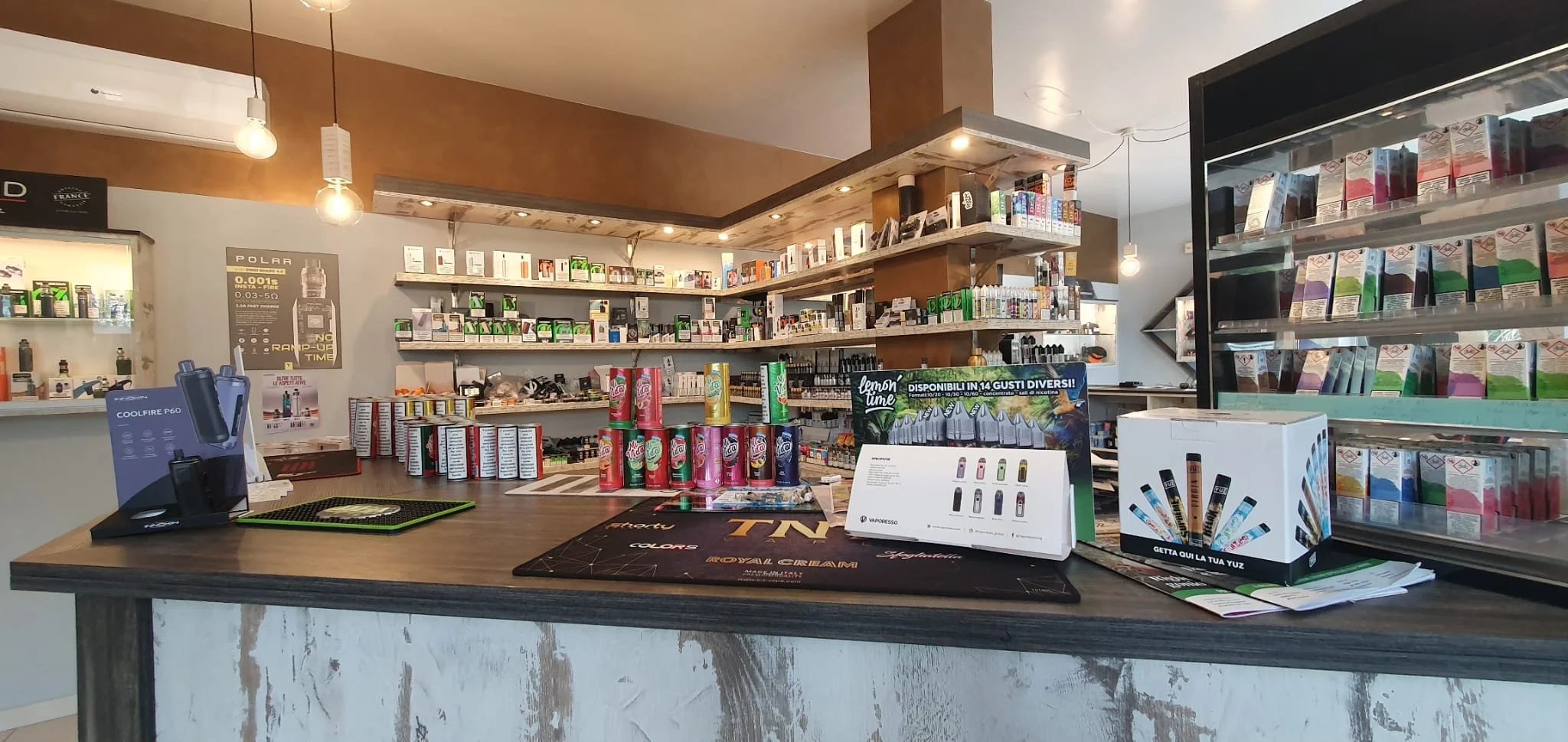 Interno di un negozio con scaffali pieni di prodotti tra cui vaporizzatori, snack e accessori, con un bancone in primo piano e luci calde sul soffitto.