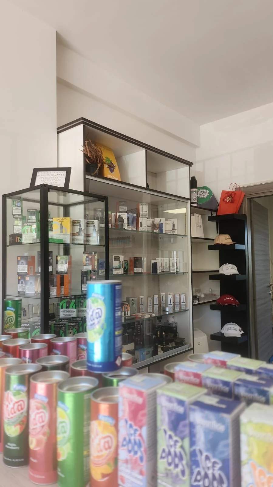 Interno di un negozio con scaffali pieni di prodotti, cappellini esposti su una mensola e un display con scatole e lattine in primo piano.