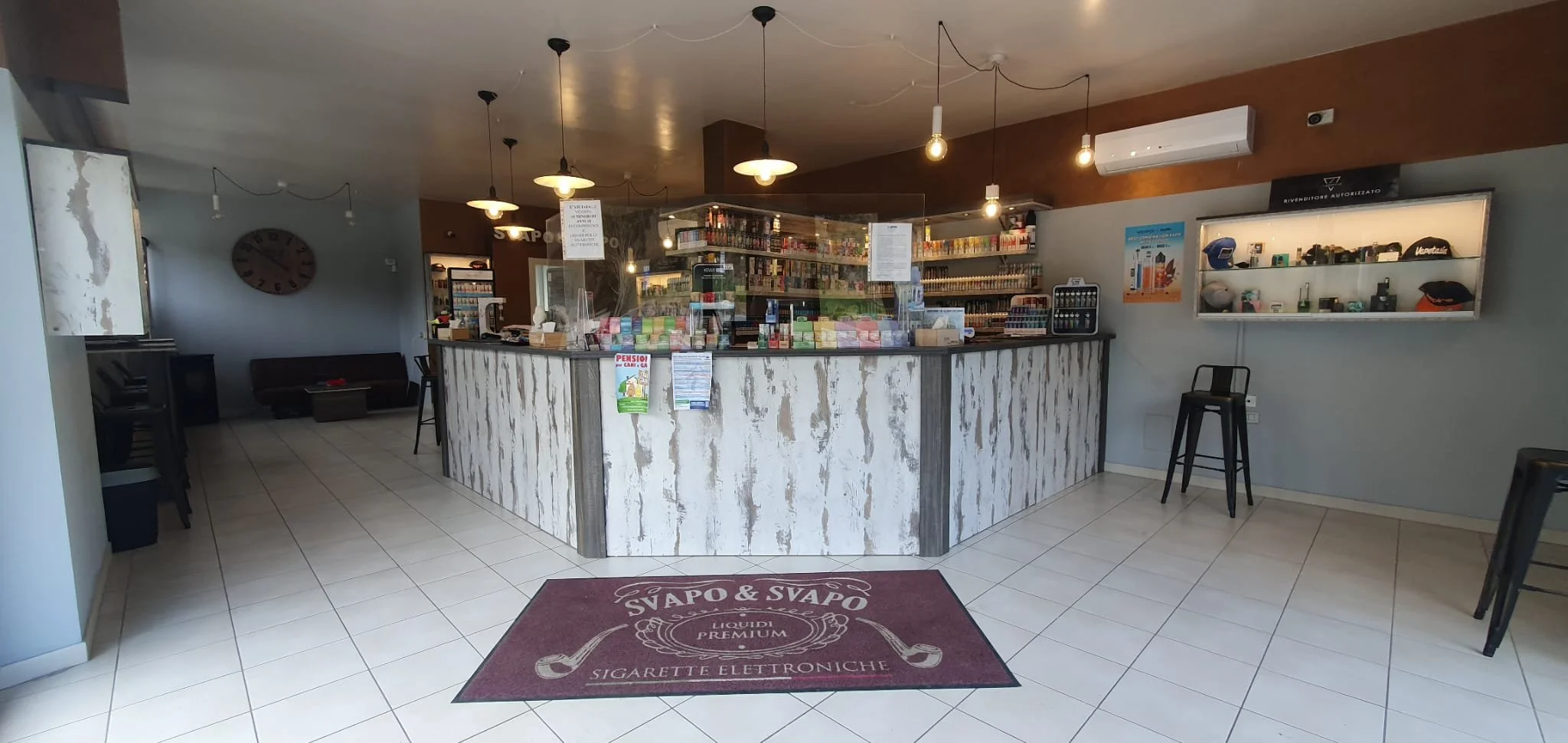 Interno di un negozio con bancone centrale, scaffali pieni di prodotti, sedie e un orologio sulla parete. Tappetino con scritta 'Svapo & Svapo' davanti al bancone.