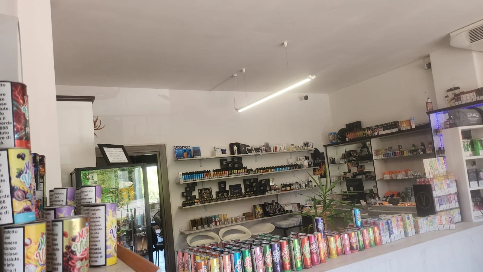 Interno di un negozio con scaffali pieni di prodotti, bancone con lattine colorate in primo piano, pareti bianche, illuminazione moderna e finestra.
