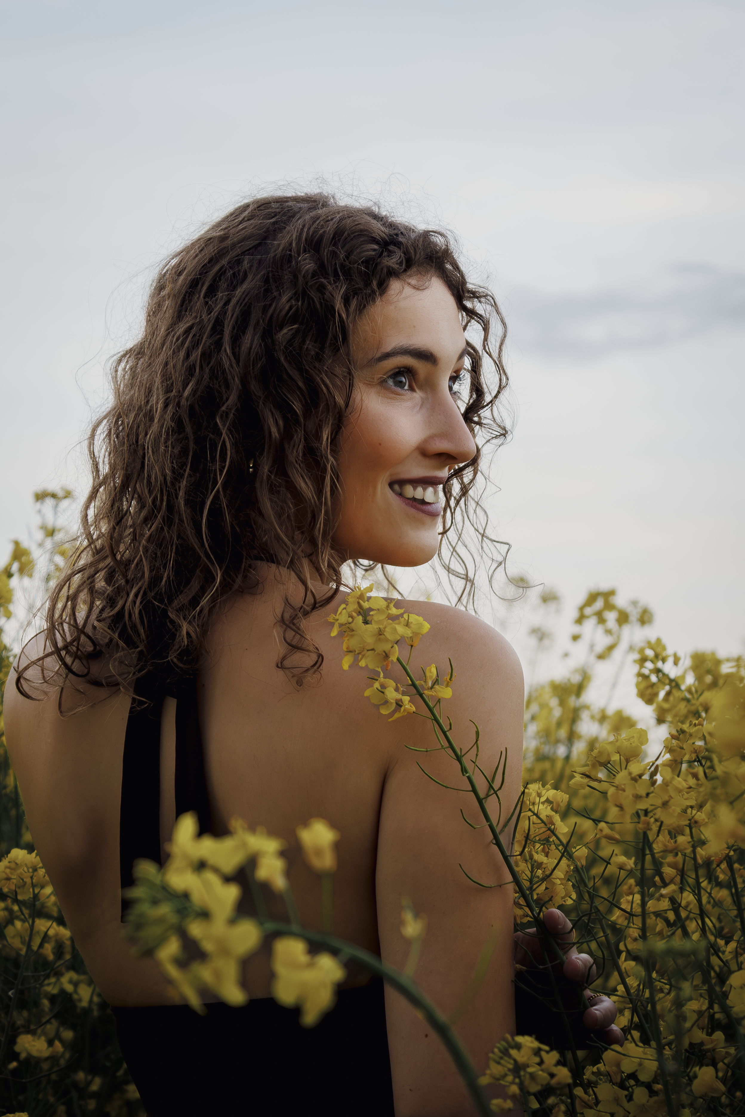 Eine junge Frau mit lockigen braunen Haaren steht in einem Feld voller gelber Blumen und lächelt, während sie nach rechts schaut, bei bewölktem Himmel.