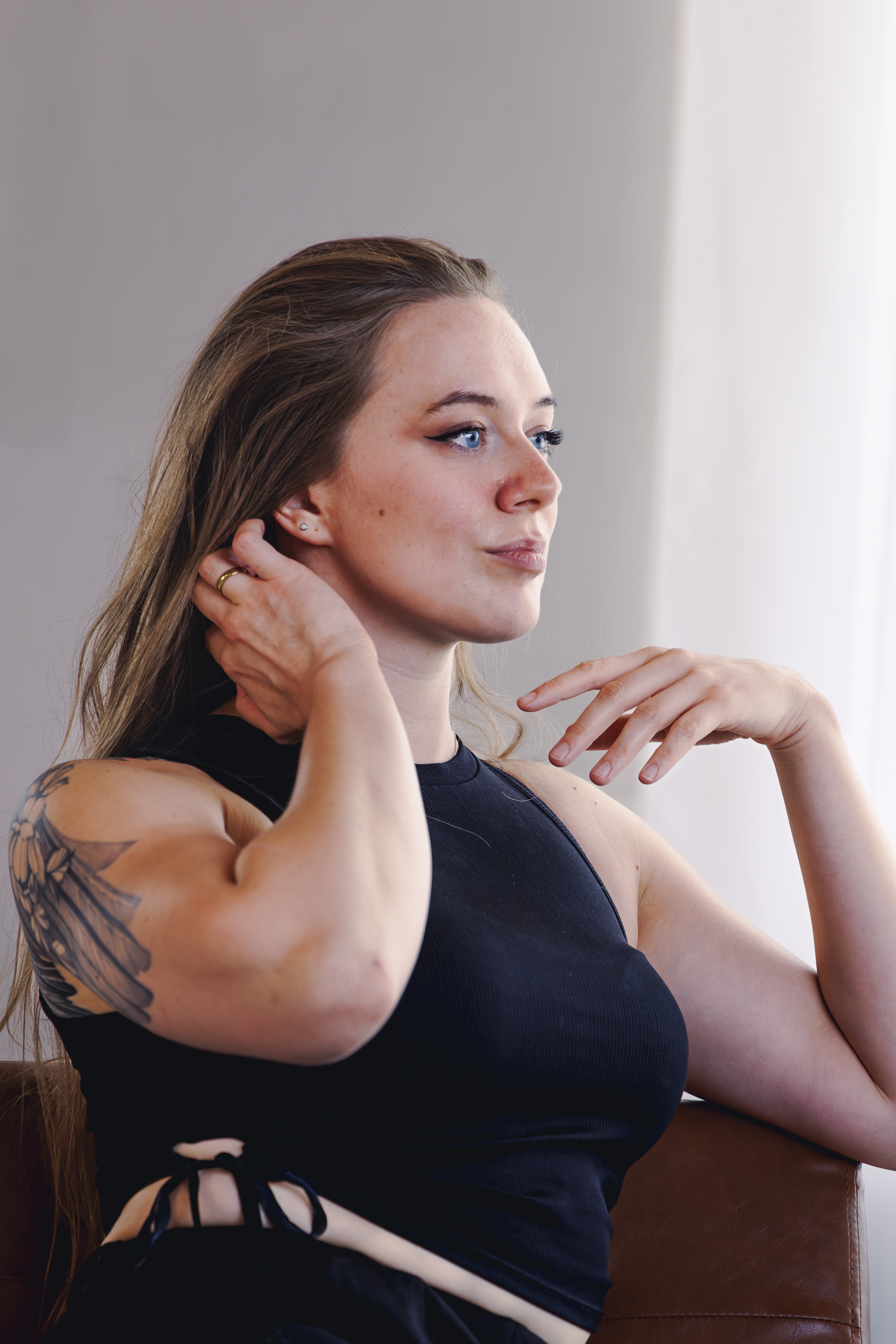 Junge Frau mit langem braunen Haar, blauen Augen, tätowiertem Arm, trägt eine schwarze Top und sitzt auf einem braunen Sofa bei natürlichem Licht.