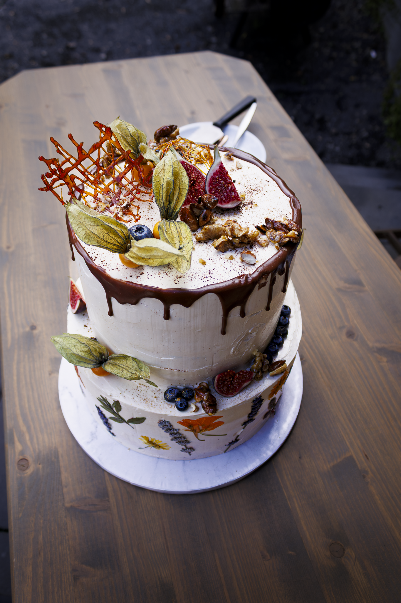 Zweistöckige Hochzeitstorte mit Ganache-Glasur, Dekorationen aus Früchten, Nüssen, Blättern und Zuckerrochen, auf einem Holztisch.