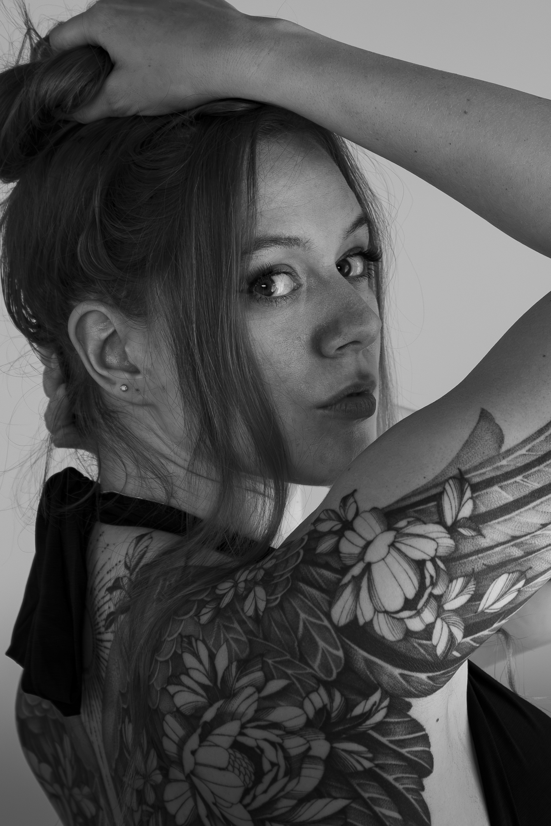 Schwarz-weiß-Fotografie eines jungen Mädchens mit langen Haaren, das eine Hand im Haar hat und den Blick Richtung Kamera richtet, mit auffälligem Tattoo auf dem Arm.