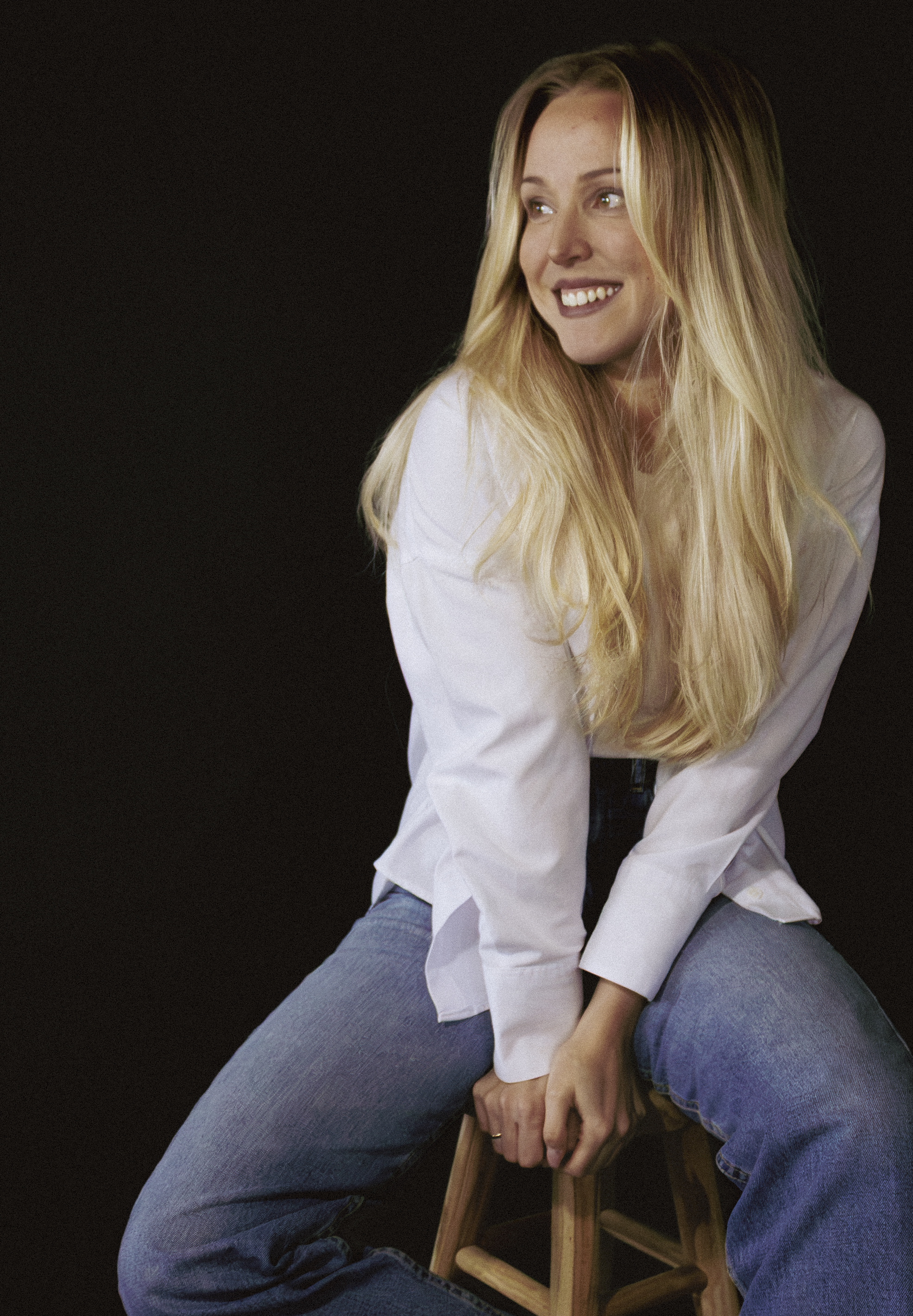 Junge Frau mit blonden Haaren, weißem Hemd und blauen Jeans, sitzend auf einem Holzstuhl vor schwarzem Hintergrund, lächelnd nach rechts blickend.