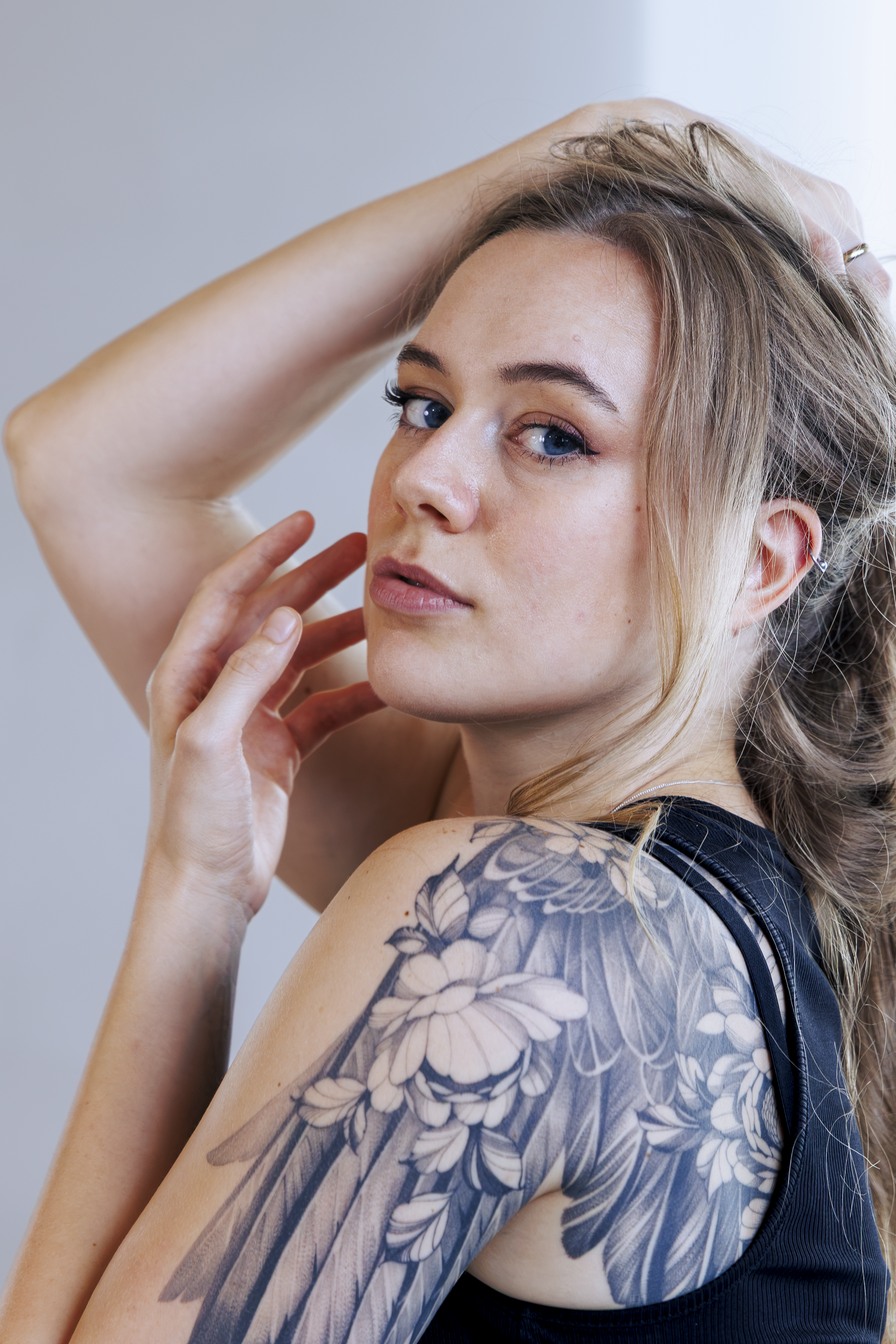 Junge Frau mit Tattoo, blonden Haaren, blauen Augen, schwarzem Top und silberner Nase, macht eine Pose mit ihrer Hand auf dem Kopf.
