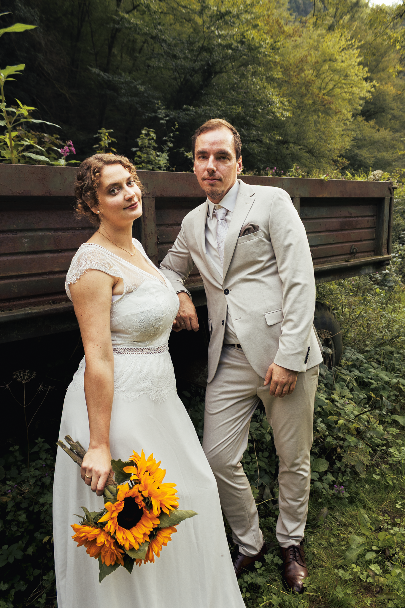 Ein Brautpaar posiert im Grünen, die Frau trägt ein weißes Hochzeitskleid und hält einen Sonnenblumenstrauß, der Mann trägt einen hellen Anzug mit Krawatte, im Hintergrund Bäume und ein altes Eisenauto.