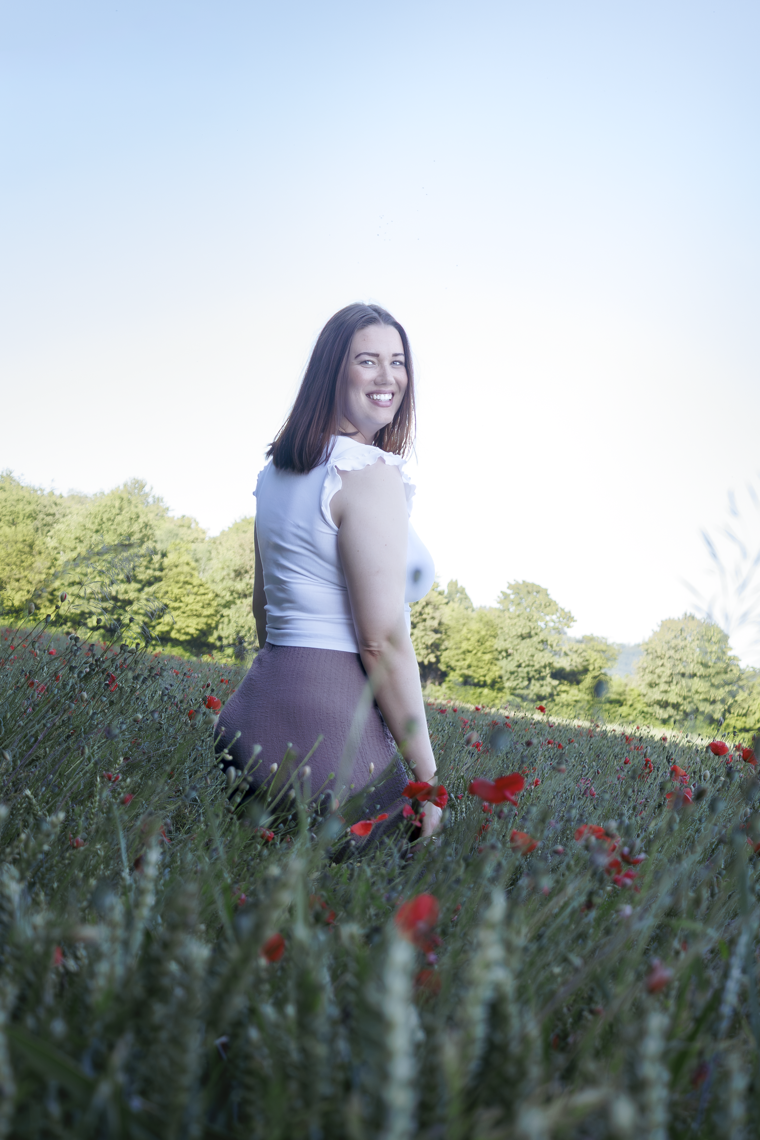 Junge Frau im Feld mit roten Mohnblumen, grünen Bäumen im Hintergrund, blauer Himmel.